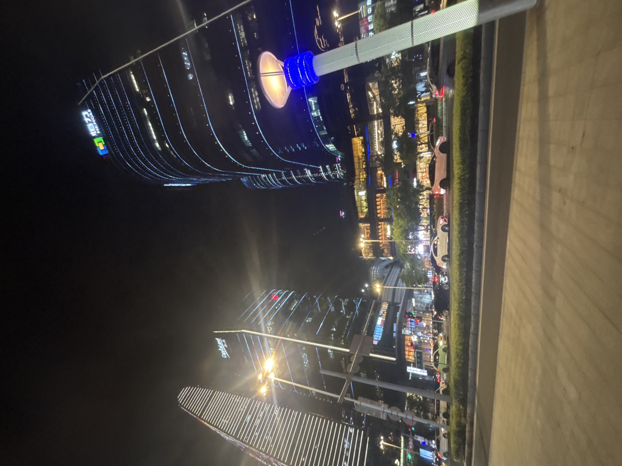 The image depicts a nighttime scene in the city of Suzhou Industrial Park, China. In the foreground, a paved walkway runs alongside a road with several cars driving by. The road is bordered by a green median strip.
In the background, towering modern skyscrapers dominate the skyline. One prominent building on the right is illuminated with blue lines and displays a logo with the text "HENGLE". To its left, another skyscraper is lit with white horizontal lines. Further to the left, a building with the "Capitaland" logo is visible, also adorned with blue lighting. These buildings are surrounded by other illuminated structures, suggesting a bustling commercial or business district. Streetlights are also visible, casting a warm glow on the surroundings. The overall impression is one of a vibrant and modern urban environment at night.