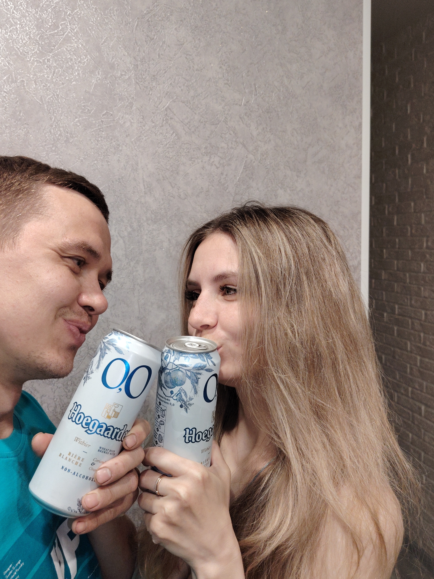 This indoor portrait features a man and a woman sharing a lighthearted moment, each holding a can of non-alcoholic beer to their lips. The man, on the left with short dark hair and stubble, wears a blue t-shirt and looks at the woman with a gentle smile. The woman, with long blonde hair, smiles faintly as she holds her can. They are positioned closely, with their faces turned towards each other, separated by the cans. Both individuals wear rings on their fingers; the woman has a simple band on her left ring finger, and the man has a ring on his right hand.
The background consists of two different textured walls: a light gray or beige patterned wall on the left and a darker, possibly brick-patterned wall on the right. A clear vertical line separates the two wall textures. The lighting is even, suggesting an artificial light source or soft natural light.
Visible text on the cans includes "0,0", "Hoegaarden", "Witbier", "BIÈRE BLANCHE", "WHEAT BEER BREWED WITH", "Coriander & Orange peel", and "NON-ALCOHOLIC". Russian text such as "Правильно" and "0,0" is also present on the woman's can. Additional text from the man's shirt mentions "Designer", "Gunter Grange", and "Foundry". The scene conveys a sense of relaxed intimacy and shared enjoyment in Chaikovsky, Russia.