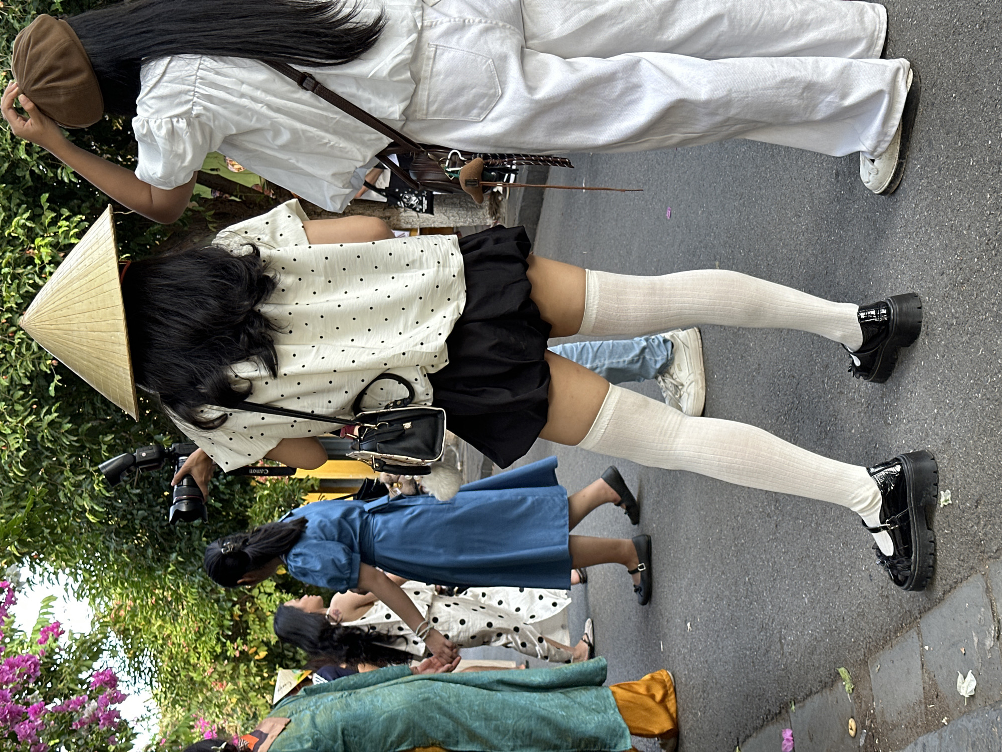 In Hội An Ward, Vietnam, a group of individuals are captured in a street scene. In the foreground, a person wearing a traditional conical hat and a white polka-dot blouse with a black skirt stands with their back to the camera. They are also wearing white thigh-high socks and black platform Mary Jane shoes. To their right, another individual in a white puff-sleeved top and white trousers is visible from the back, accessorized with a brown beret and a brown crossbody bag. On the left, a person in a green and orange outfit walks away from the camera. Behind them, a person in a blue dress is walking, and another person in a white polka-dot dress with a black pattern is also visible. The ground is paved with asphalt, and foliage from trees can be seen in the background. A camera on a tripod is being held by the person in the conical hat.
