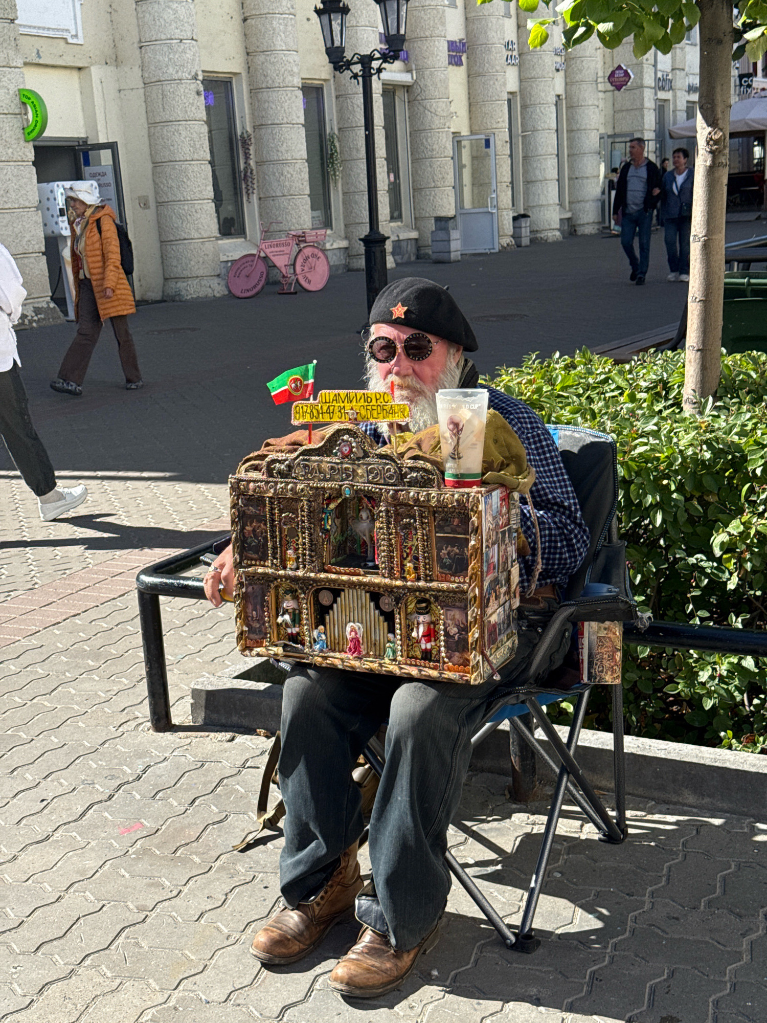 An elderly man with a white beard and sunglasses, wearing a black beret with a red star, sits in a folding chair on a city sidewalk in Kazan, Russia, near a leafy green bush. He holds a small, ornate, multi-level music box adorned with miniature figures and framed pictures. A clear plastic cup with a beverage sits on top of the music box, next to a small green cloth. A flag, appearing to be the flag of Chechnya, is planted on the music box. The man wears a blue and black checkered shirt and dark jeans, and brown leather boots. The background shows a building with large columns, two lampposts, and people walking in the distance. A pink bicycle with decorative wheels is visible on the sidewalk further down. The ground is paved with interlocking grey bricks.