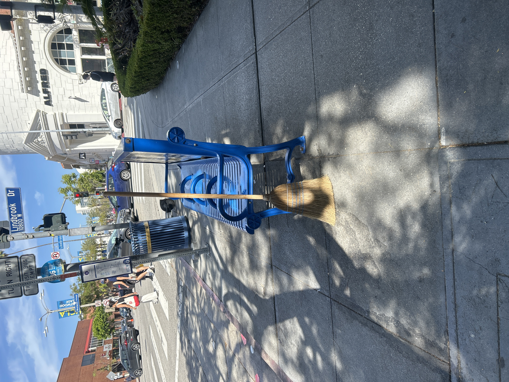 This is an outdoor shot of a blue park bench with a broom lying across it, located on a sidewalk in Westwood Village, Los Angeles. The street sign indicates the location as Lindbrook Drive, and there are signs for Westwood Village nearby. In the background, there are people standing at a bus stop. Beyond that, we can see traffic and buildings typical of an urban environment. The weather is sunny, as indicated by the shadows on the sidewalk.