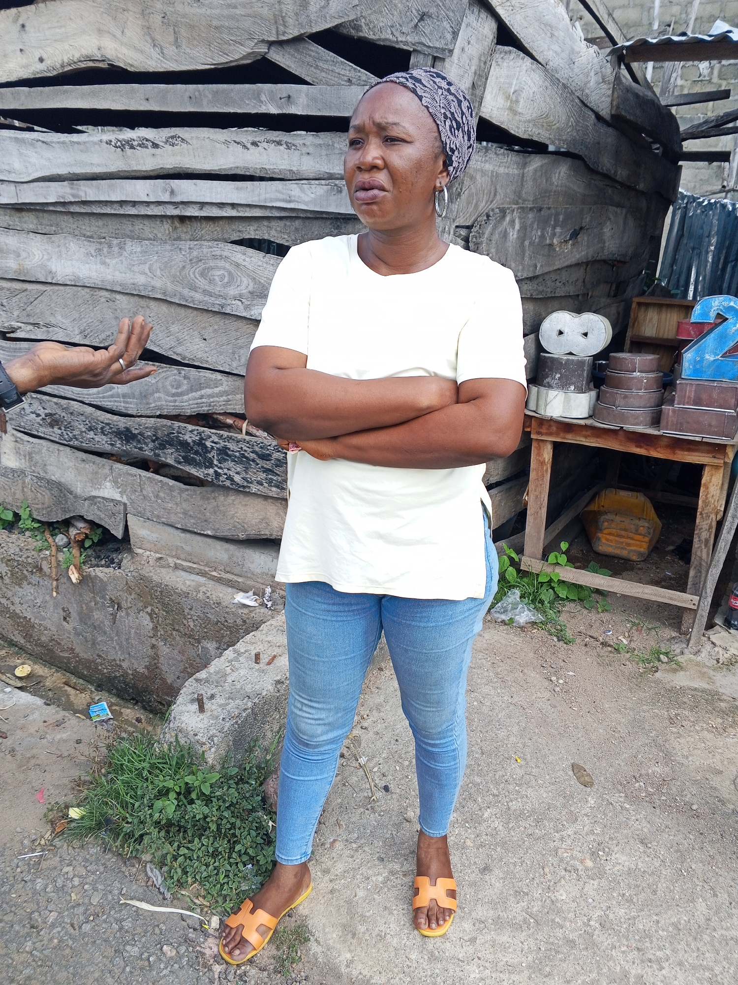 In Zango-ndaji, Nigeria, a woman stands with her arms crossed, wearing a white t-shirt, blue jeans, and orange sandals. She is positioned in front of a structure made of weathered wooden planks. To her right, a makeshift wooden table displays various objects, including stacked circular molds and a blue numeral "2." Behind her, a rough, concrete channel is visible with some overgrown vegetation. The ground is a mix of dirt and gravel. A person's hand, wearing a watch and a ring, is partially visible on the left side of the frame, gesturing towards the woman. The scene suggests an outdoor market or workshop environment.