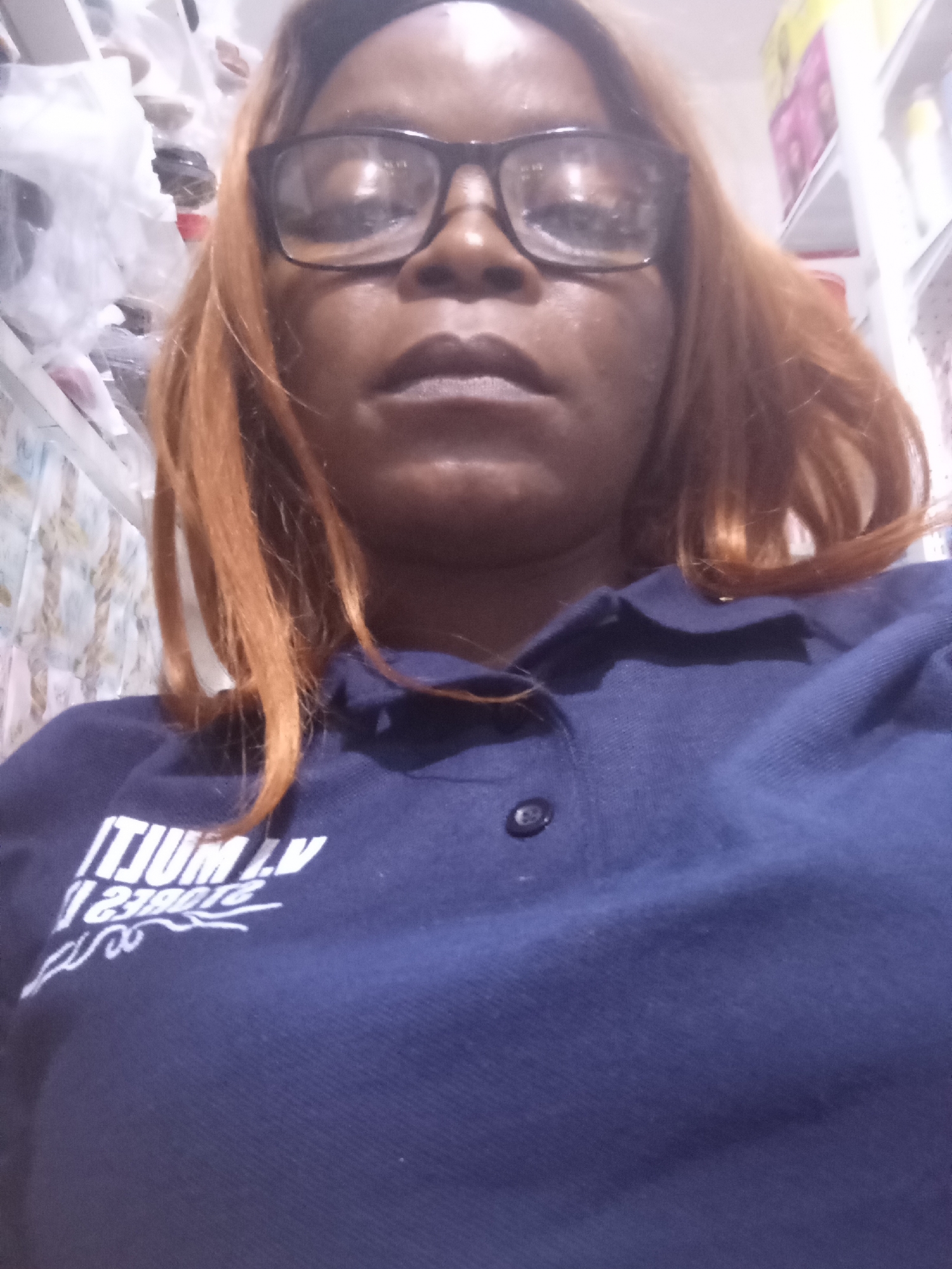 A woman with auburn hair and glasses looks directly at the camera. She is wearing a dark blue polo shirt with white lettering that reads "ITIUM LV 238018". The background is blurred but appears to be a retail environment with shelves of items, some covered in plastic. The lighting is artificial and the image has a slightly grainy quality. The overall scene suggests an informal selfie taken indoors.