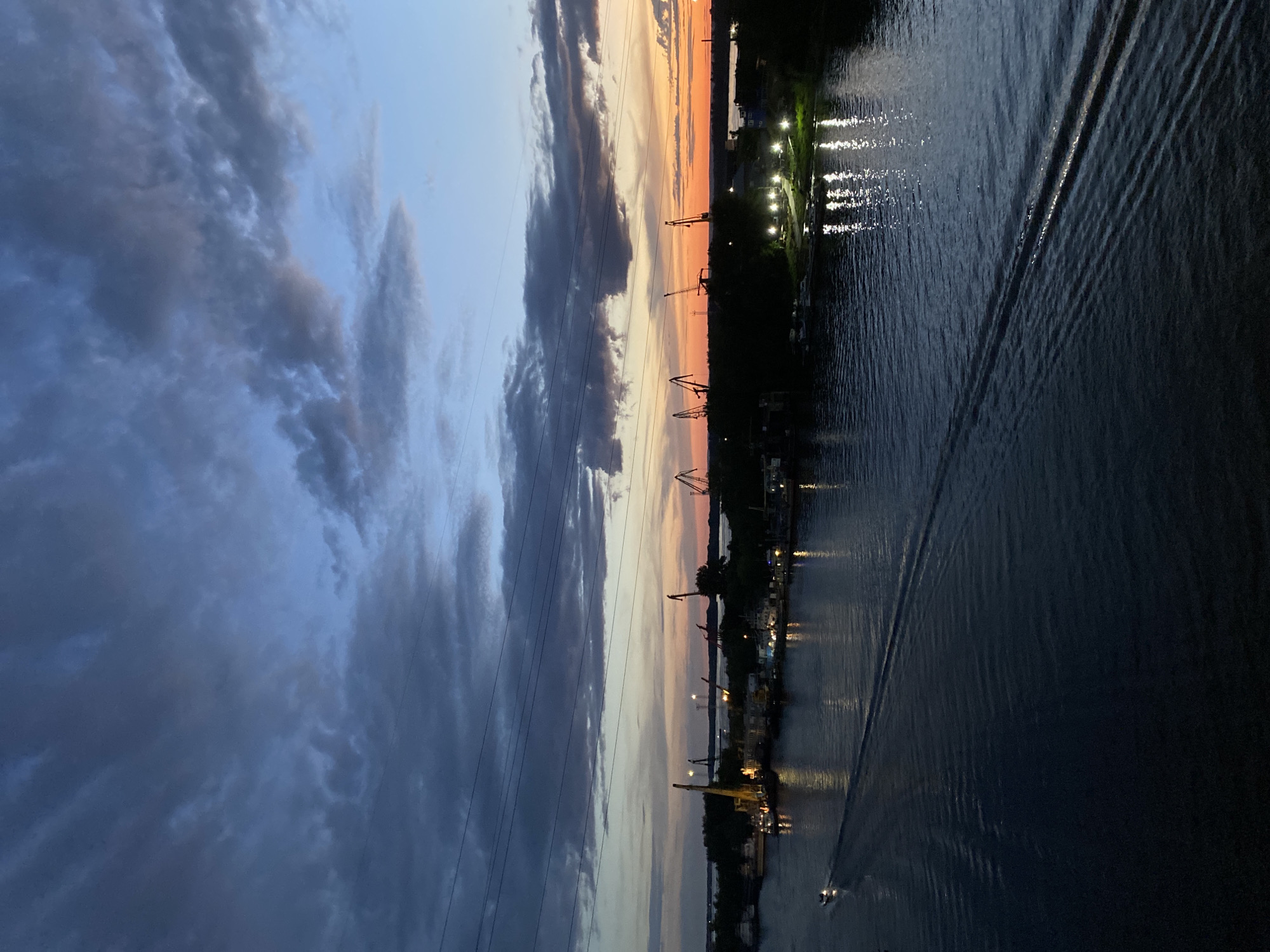 The image depicts a twilight scene over a body of water, likely a river or canal, with an industrial shoreline. The sky is a dramatic mix of deep blue and grey clouds on the left, transitioning to a soft orange and pink sunset on the right, hinting at the end of the day.
Along the water's edge, silhouetted against the fading light, are several tall cranes, characteristic of a port or industrial area. Lights are visible along the shoreline, reflecting on the water's surface, creating a shimmering effect. The water itself is dark and calm, with visible wakes trailing behind a small boat that is moving across the frame. A larger boat or barge appears to be docked or moving slowly near the shore, marked by lights.
The overall mood is serene yet industrial, with the natural beauty of the sunset contrasting with the man-made structures. The presence of power lines running across the sky adds to the sense of an inhabited and operational area. The time of day is clearly dusk or early evening, as indicated by the low angle of the sun and the artificial lights illuminating the scene. The weather appears to be clear apart from scattered clouds. While the specific location is not definitively identifiable from visual cues alone, it is described as Novokuybyshevsk, Russia. No text is visible in the image.