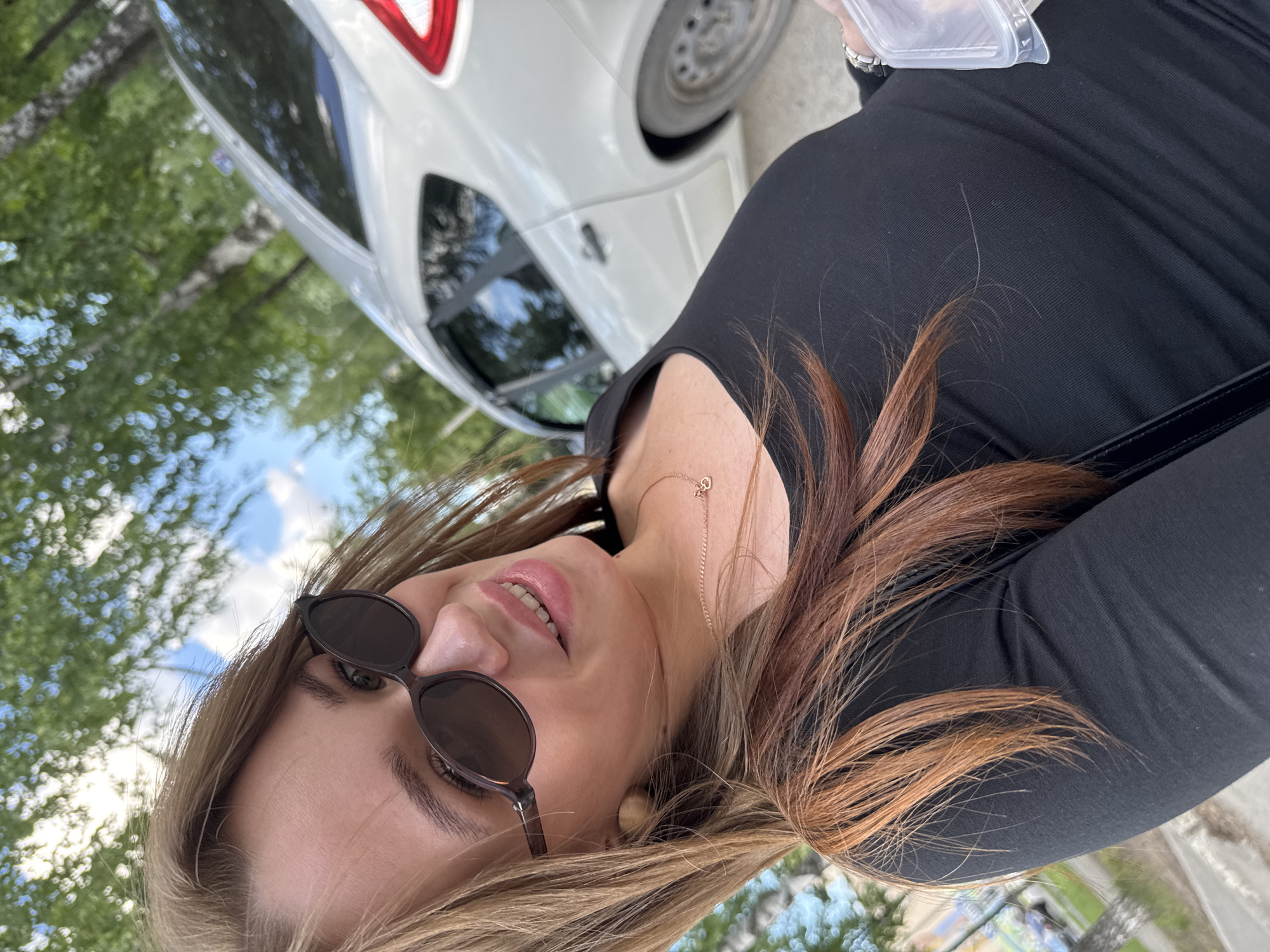 A young woman with long, reddish-brown hair and fair skin takes a selfie outdoors. She is wearing sunglasses and a black long-sleeved shirt. A gold necklace with a small pendant is visible. In the background, a white car is parked on a street lined with trees. The sky is blue with some clouds, and sunlight filters through the leaves, creating a dappled effect. The setting appears to be a residential area or a park in Chaikovsky, Russia, given the presence of birch trees and the characteristic architecture of a Russian city. The woman holds a clear plastic container in her right hand.