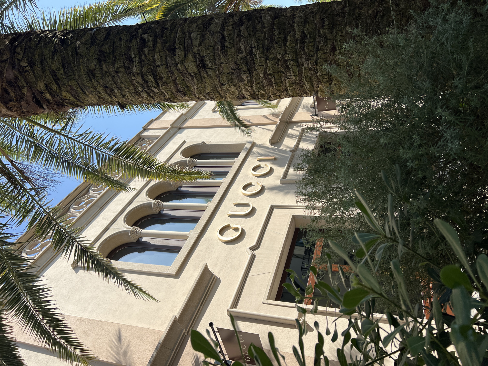 Captured from a low angle, the image features the façade of a building, with the word "GUCCI" vertically displayed in golden letters. The architectural style suggests a luxury retail location. Palm trees frame the top and left of the shot, adding a tropical element. The presence of lush green foliage on the right further accentuates the outdoor setting. Given the provided location of La Roca del Vallès, Spain, this is likely the Gucci store located in the La Roca Village outlet shopping center.