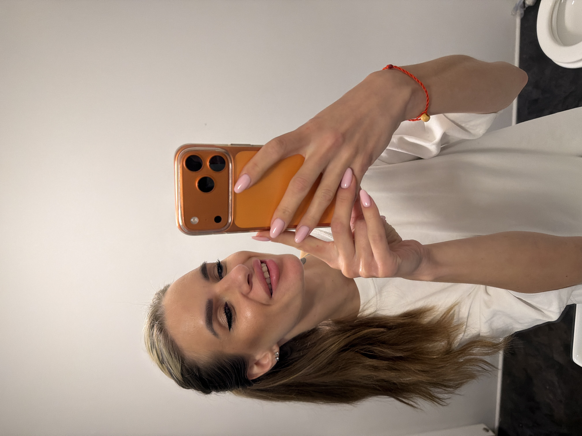 A female subject, appearing to be an adult, is depicted taking a mirror selfie indoors, in Berlin, Germany. The specific location within Berlin is not discernible from the image, which shows a private or commercial bathroom setting. The subject has long, light brown hair with blonde highlights, pulled back into a ponytail. She is wearing a white short-sleeved t-shirt. A small, clear stud earring is present in her visible left ear. She is smiling, with visible makeup including eyeliner and pink-toned lip color. She holds an orange-cased smartphone, likely an iPhone, in her right hand, displaying light pink nail polish. A red string bracelet with a single light-colored bead is worn on her right wrist. The background features plain white walls and a dark tiled floor. A white ceramic fixture, consistent with a toilet or sink, is partially visible in the lower portion of the frame.