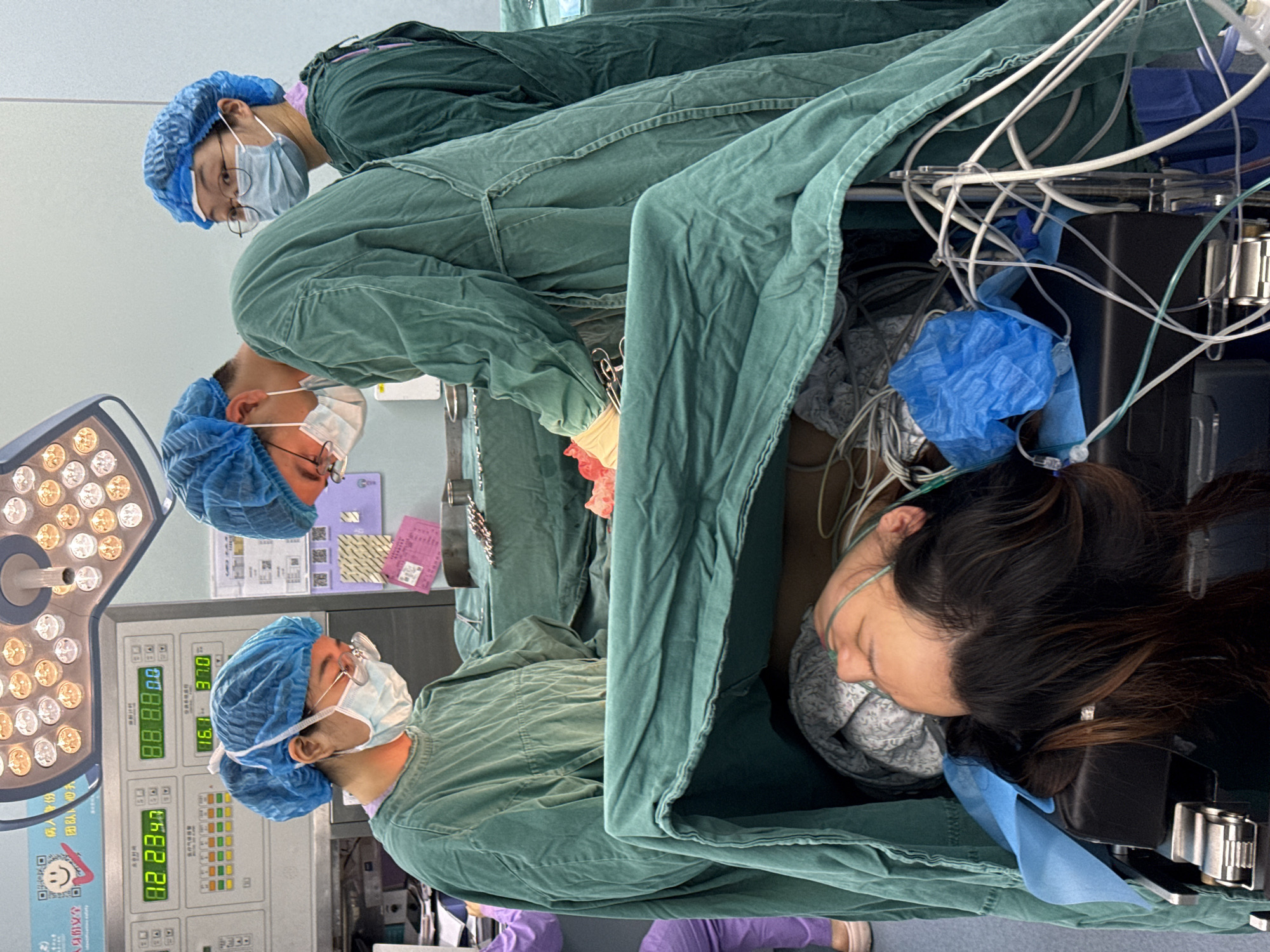 The image depicts an operating room setting. Three individuals, presumably surgeons or medical staff, are seen wearing surgical caps and masks, dressed in green surgical gowns. They are positioned around an operating table draped in green surgical sheets, partially obscuring the patient. Wires and medical equipment are visible in the background. Overhead, a surgical lamp illuminates the scene. The location is likely a hospital or surgical center, possibly within Yuzhong District, China, but there is no explicit evidence of this in the image.