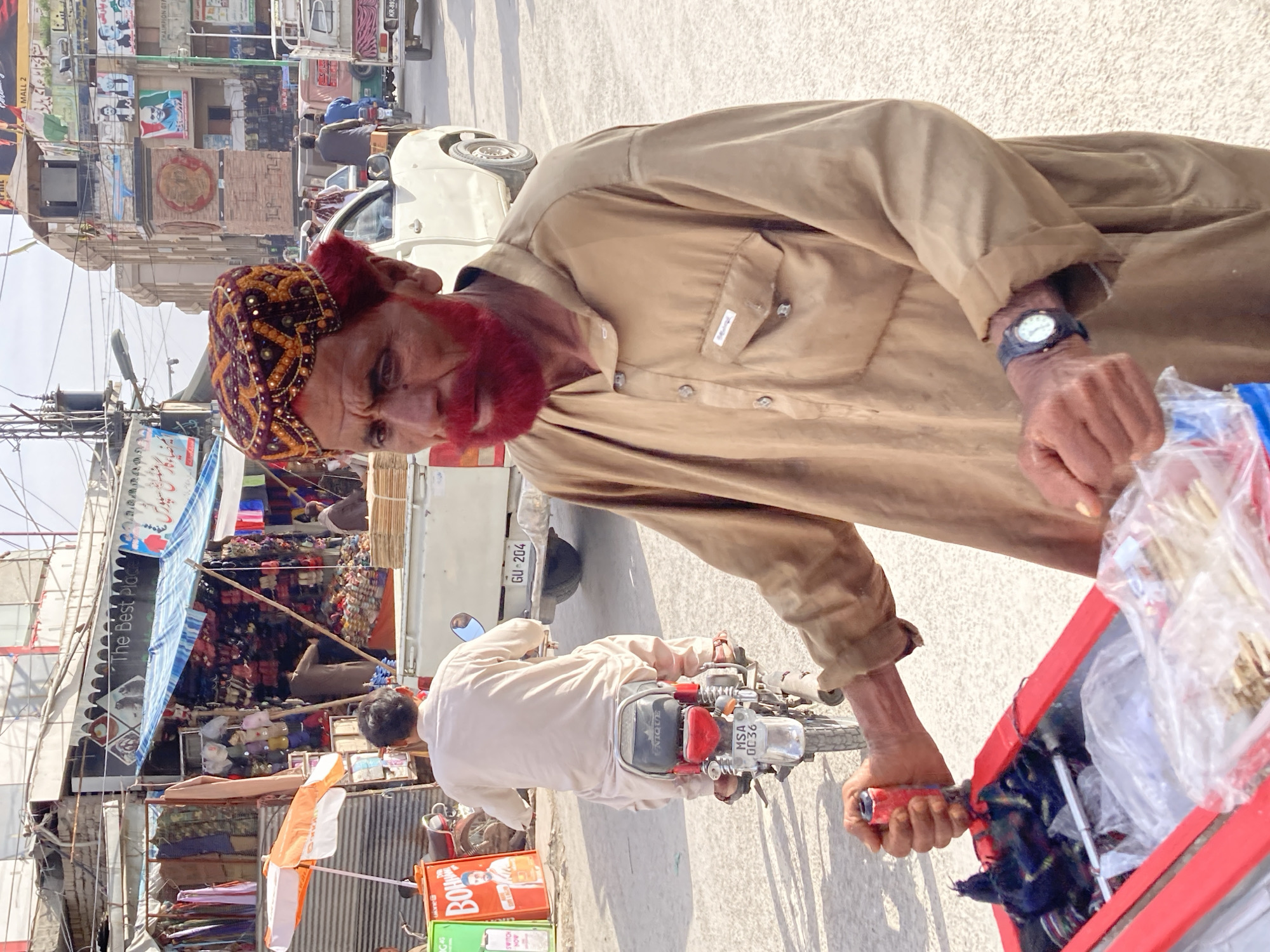 This media file captures a vibrant street scene in Quetta City Tehsil, Pakistan, under bright daylight conditions. The image is dominated by an elderly man in the foreground, presumably a street vendor, looking directly at the viewer. He wears a traditional embroidered cap with gold and purple patterns, a light tan long-sleeved shirt, and has a distinctive red-dyed beard and mustache. A watch is visible on his left wrist as he holds onto a bright red cart or display stand that contains various items, possibly small tools or trinkets, packaged in clear plastic bags.
Behind the vendor, the street is bustling with activity. A man on a motorcycle, wearing light traditional attire, is riding past; the motorcycle's license plate reads "MSA 0C36" and "UNIQUE" is visible on the seat. A white car with license plate "GU-204" is parked further back. The background features a lively commercial area with multi-story buildings housing numerous shops displaying colorful merchandise like fabrics and shoes. A light truck with license plate "KR-893" is also visible.
The scene is typical of a busy market street, characterized by overhead wires, awnings, and numerous signs. Visible text includes English phrases like "The Best Place," "MALL 2," "CHAIMPION GT," and "SWITCH NOW" on a green box, along with "BOH" and "ulonel" on a red box. Several banners with Urdu script and political posters featuring portraits and symbols (such as a green flag with a star and crescent) are also present. The strong shadows suggest a midday setting, and the overall atmosphere is one of daily commerce and urban life in Pakistan.