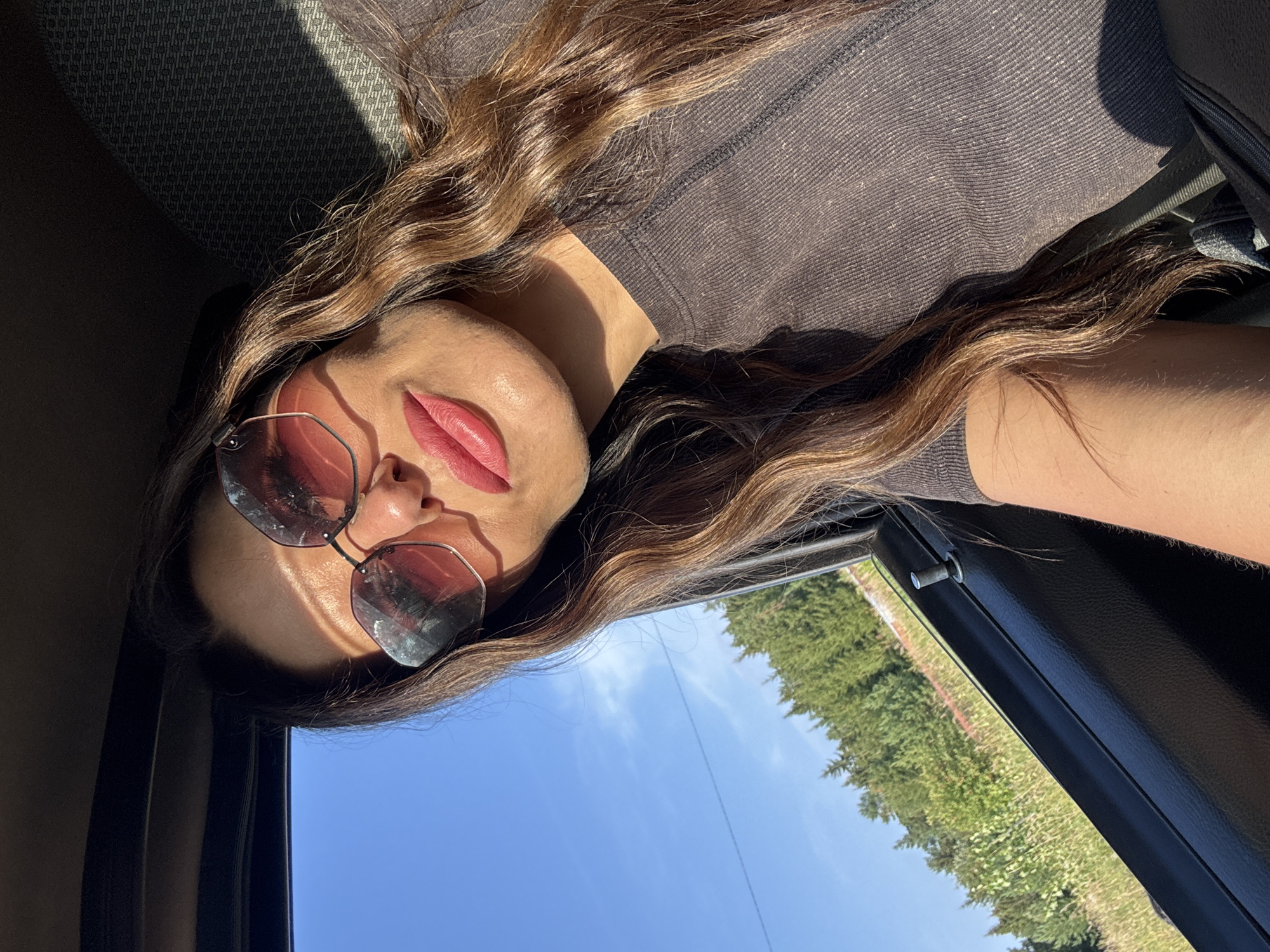 A woman with long, wavy brown hair and sunglasses with rose-tinted lenses poses for a selfie inside a vehicle. Her lips are painted a vibrant pink. Sunlight casts a shadow across her forehead. The background shows a bright blue sky and a dense forest with green trees, suggesting an outdoor setting, possibly near Olkhovka, Russia, given the description. The interior of the car is visible, with a black door panel and a dark-colored seat.