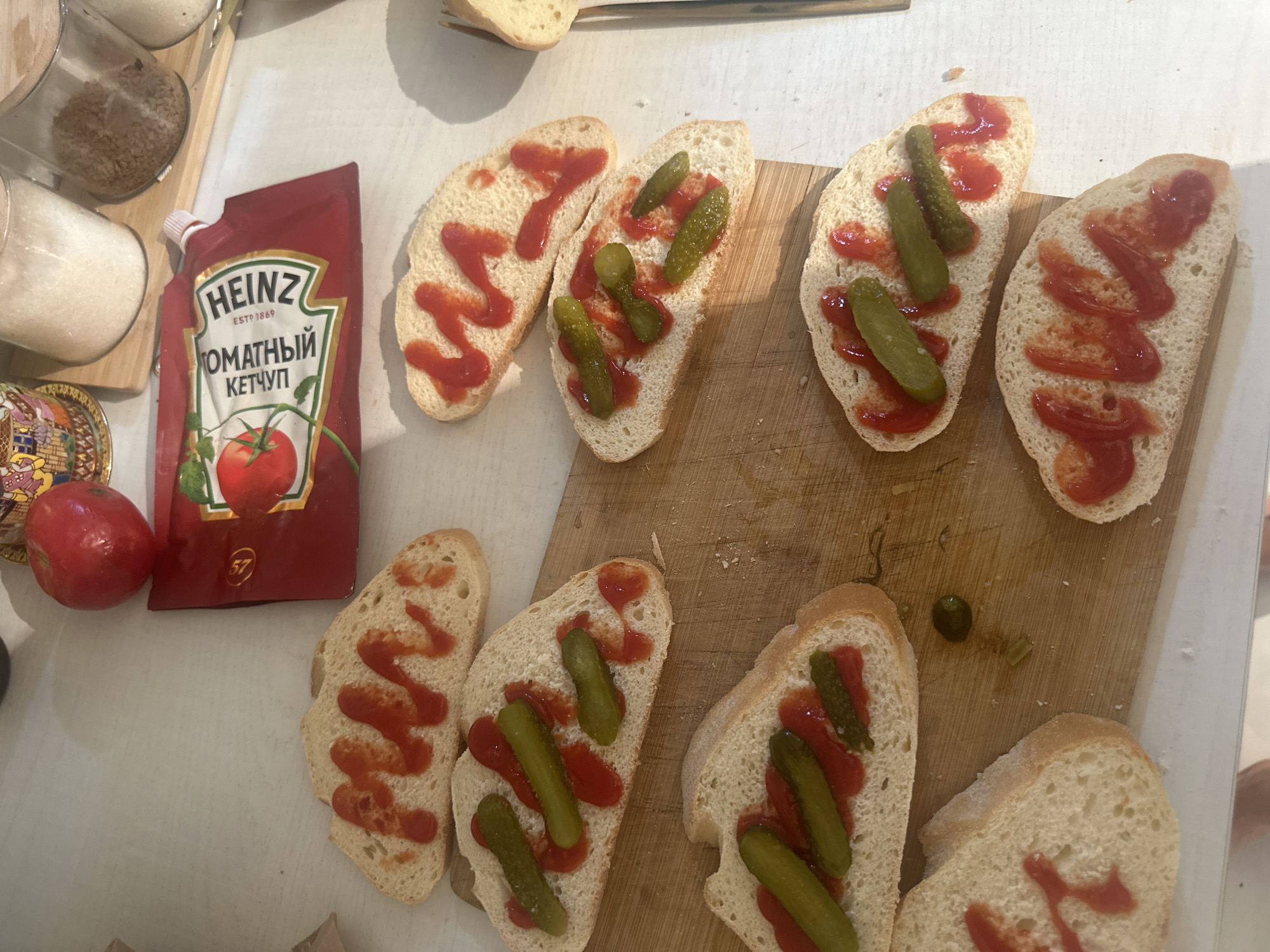 The scene is a close-up, overhead shot of a kitchen counter in Khimki, Russia. Several slices of white bread are arranged on a light-colored wooden cutting board. Most of the bread slices are topped with ketchup and sliced pickles. A red Heinz ketchup pouch with Cyrillic text is placed next to a tomato. Several jars containing grains or sugar are in the upper left of the frame. The lighting is bright and appears to be natural.