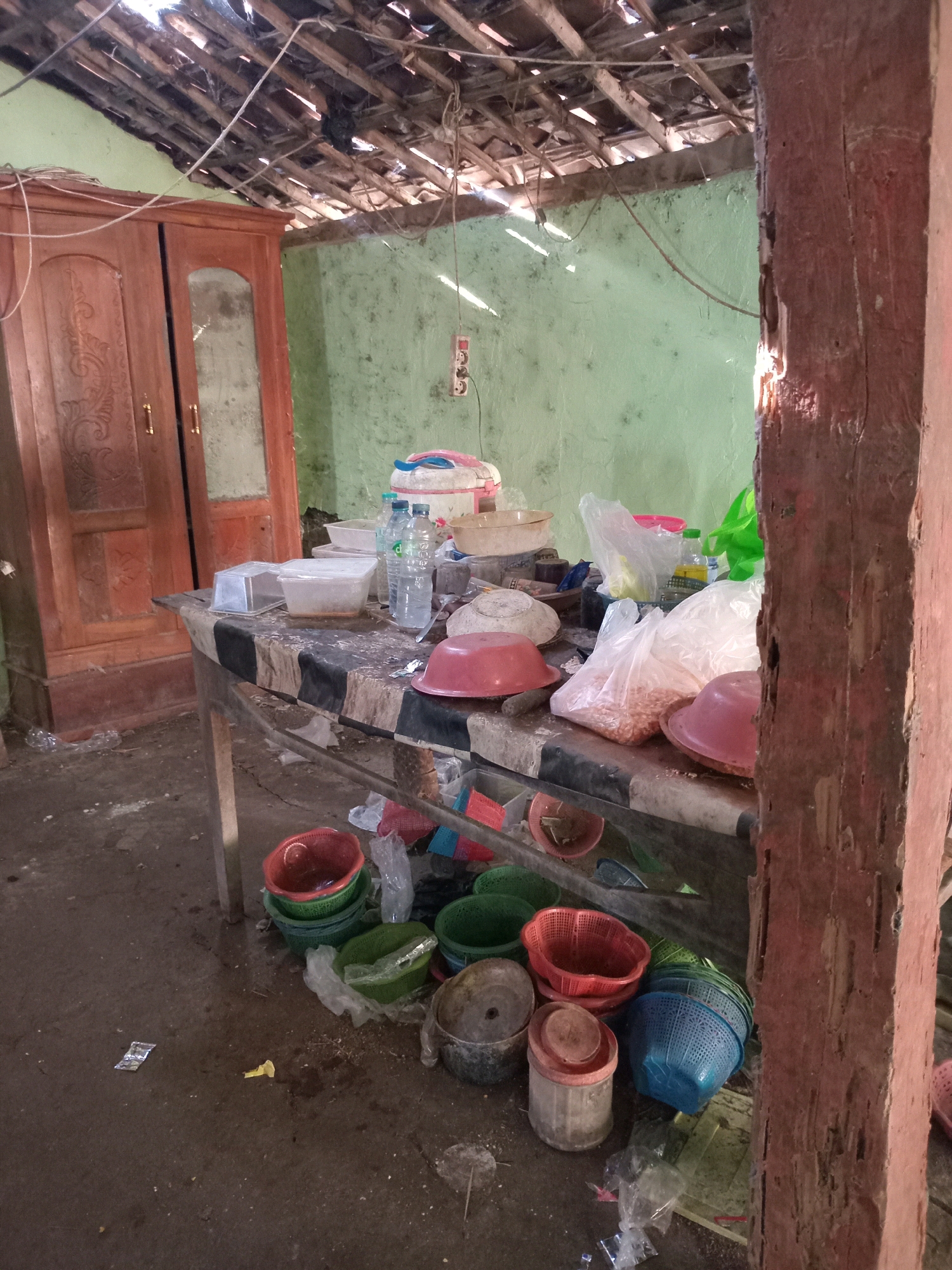 The image depicts an interior scene, likely a humble dwelling or storage area in Mojorayung, Indonesia. A rough wooden table, covered with a checkered cloth, dominates the foreground. On the table are various items, including plastic containers, bottles of water, and bags of what appear to be dry goods like peanuts. Several red and pink plastic bowls are scattered across the table and on the floor.
Beneath the table, more colorful plastic baskets are piled, some containing other items or appearing empty. The floor is concrete and appears somewhat damp and dirty, with scattered debris and a few plastic wrappers.
The walls of the room are painted a pale green and show signs of wear. Above, the ceiling is constructed of wood beams and what seems to be roofing material, with gaps allowing light to filter through. A wooden wardrobe with ornate carvings stands against the left wall. A thick wooden post, rough and weathered, is prominent on the right side of the frame, suggesting a structural element of the building. The overall impression is one of a functional but unrefined living or storage space.