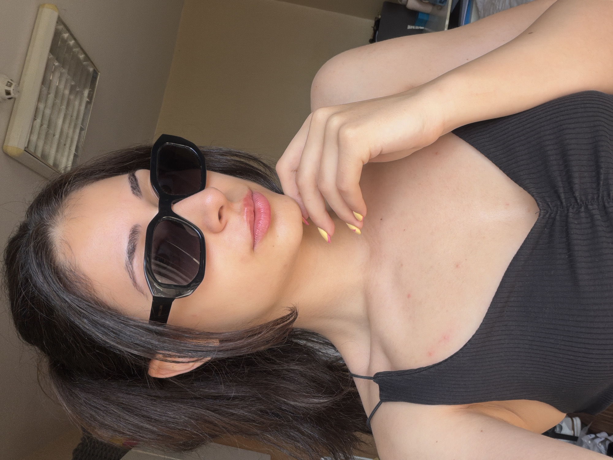 A woman is lying on her side. She is wearing sunglasses and a black top. Her hand is resting on her chin. The location cannot be determined from this image.