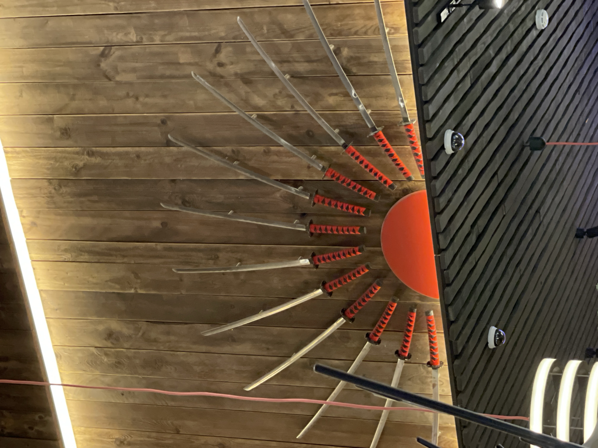 The image shows a wall decorated with an arrangement of swords, resembling a rising sun. The swords have red and black patterned handles and are mounted on a wooden plank wall. A bright light source is positioned above the swords. In the foreground, a black pole and a pink wire are visible. Below the wall, a dark ceiling with angled wooden slats can be seen. Two surveillance cameras are mounted on the ceiling, along with a circular light fixture. The scene appears to be indoors, possibly in a restaurant or commercial establishment. There are no people visible in the image. The location is identified as Moscow, Russia.