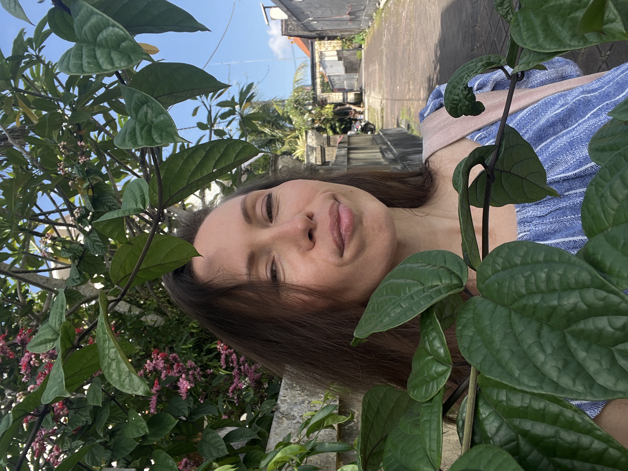 A person is seen in a medium shot, seemingly taking a selfie, in what appears to be an outdoor alleyway in North Kuta, Indonesia. The person has long brown hair and is partially obscured by leafy green vegetation on both sides of the frame. Behind the person is a narrow paved alleyway with buildings on either side, indicating a residential area. The buildings are constructed of light-colored stone or concrete, typical of the region. Tropical plants and palms are visible in the background between the buildings. The sky is partially visible and appears to be clear and blue. The person is wearing a blue and white striped top.