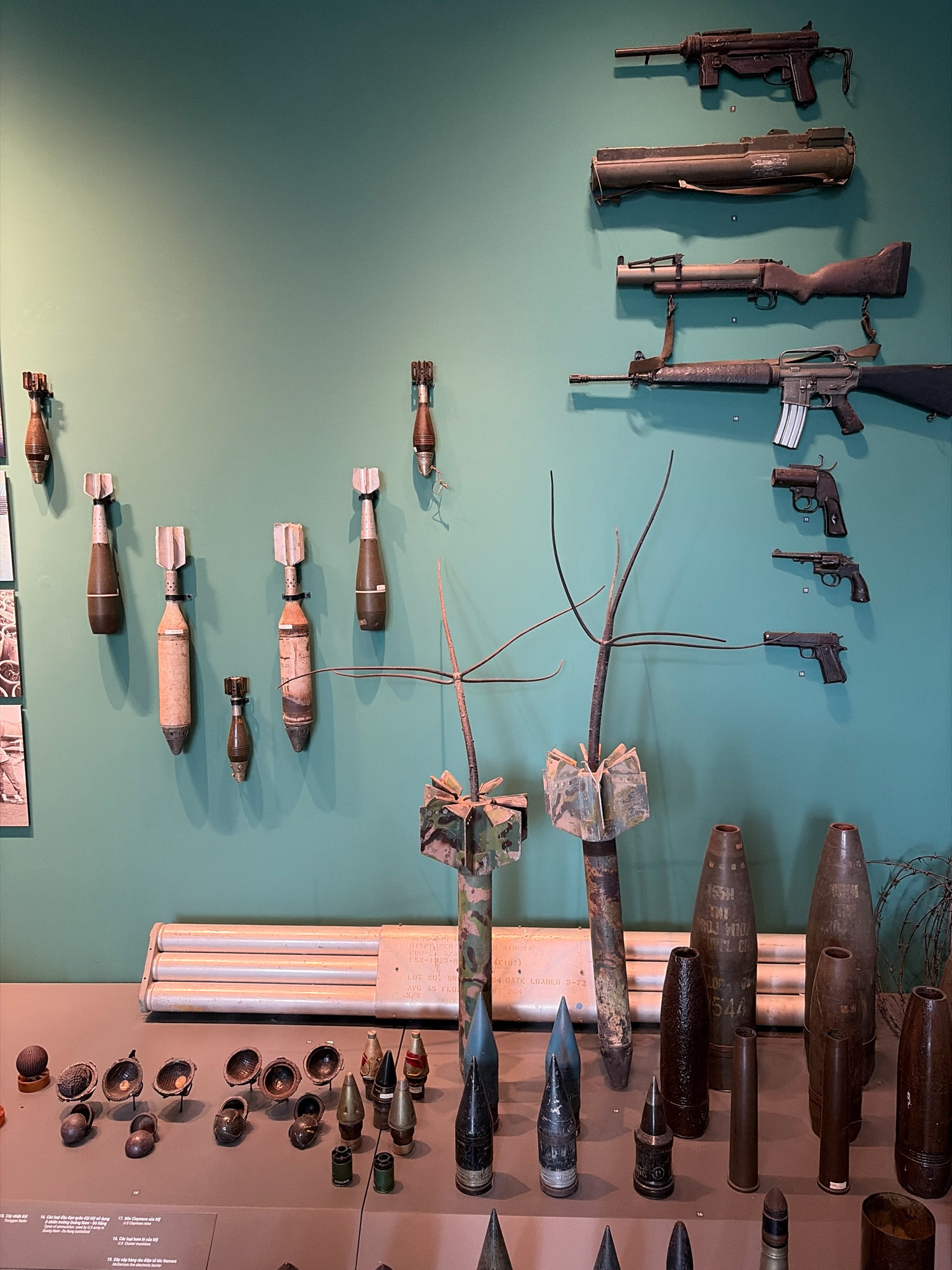 The image displays a collection of military artifacts, including firearms and ordnance, mounted on a pale green wall and laid out on a display shelf in what appears to be a museum. The firearms are positioned vertically on the wall, starting with a submachine gun at the top, followed by a rocket launcher, a rifle with a wooden stock, and an assault rifle. Below these, a series of handguns are arranged vertically, including a revolver and two semi-automatic pistols. Hanging on the wall to the left are several mortar shells, each with fins at the top.
On the display shelf in the foreground, an array of ammunition, including artillery shells and smaller projectiles, are presented. Among these are several conical-shaped objects that appear to be practice bombs or inert ordnance. Behind these, a multi-barreled projectile launcher or rocket system is visible, mounted horizontally. The background consists of the pale green wall and the display shelf.
Based on the text visible on the labels of some items on the display shelf, which includes Vietnamese characters and references to "Mỏ lao", "Cây", and "bài", and given the context of military artifacts commonly displayed in Vietnam, this scene is likely located at the Da Nang Museum of Military Zone 5 in Da Nang, Vietnam. The museum is dedicated to preserving and exhibiting the history of the military in the region. The lighting is even, suggesting an indoor, well-lit environment, typical of a museum exhibition. No people are visible in the image.
