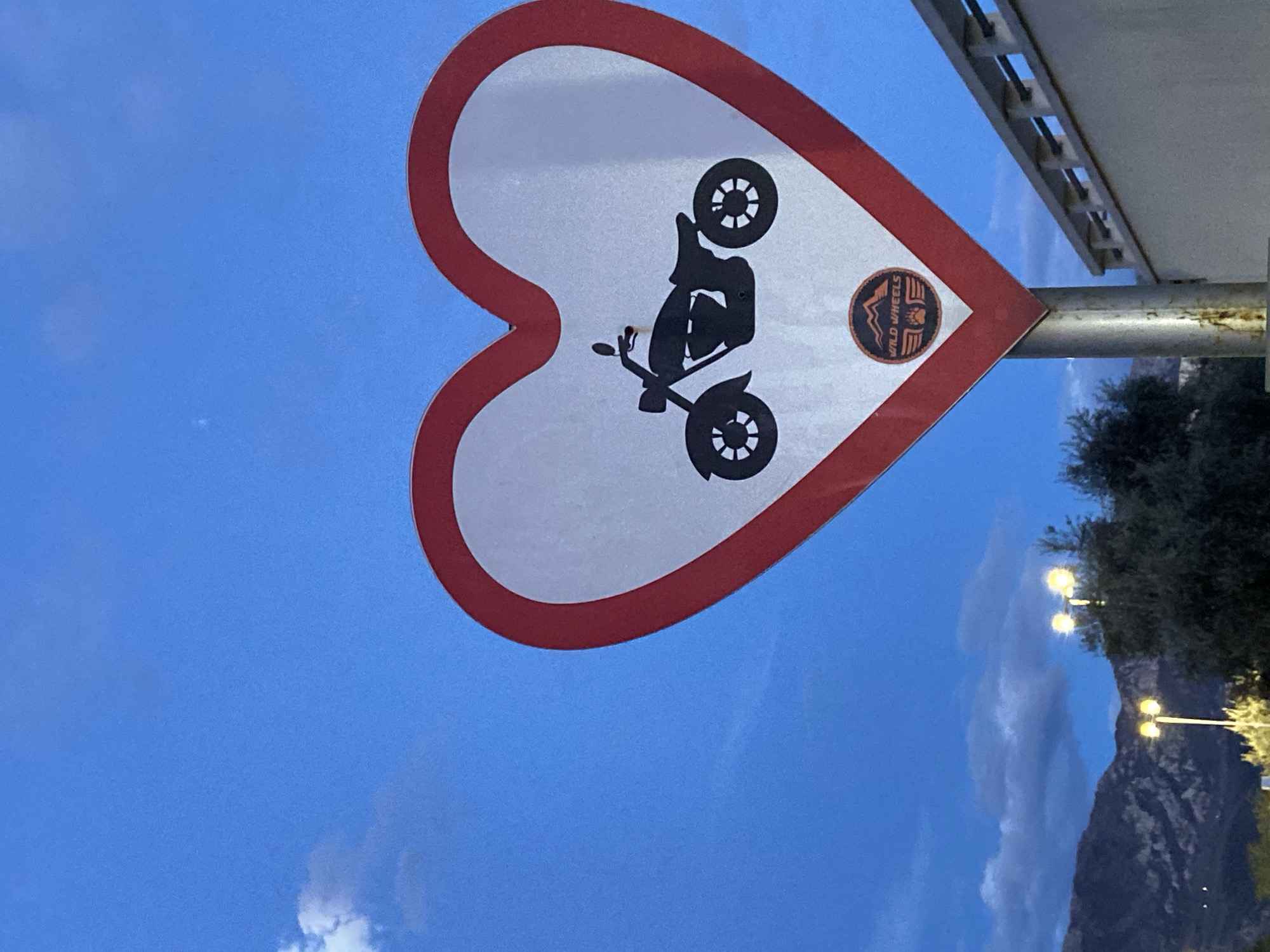 Here's a detailed description of the image:
The image shows a road sign, heart-shaped with a white background and a red border, depicting a black motorcycle silhouette and a "Wild Wheels" logo sticker. The sign is mounted on a silver pole. The background consists of a light blue sky, with some clouds. In the lower right corner, a mountain landscape with streetlights is partially visible. The location is inferred to be Boreti, Montenegro, based on the logo sticker on the sign. The sign is rotated at an angle.