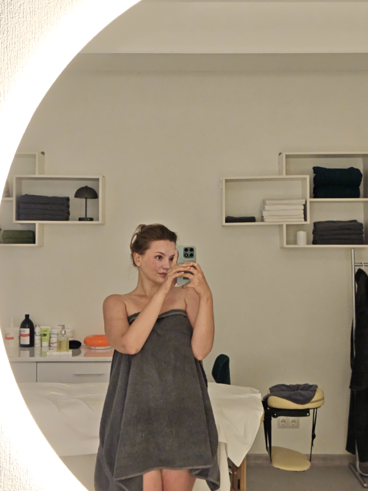 In Vinnytsia, Ukraine, a young woman, with her hair in a bun, stands in front of a large, illuminated oval mirror. She is wrapped in a dark gray towel and holds a smartphone in her right hand, taking a selfie. The background reveals a clean, well-lit room, likely a spa or treatment center, with white walls. To her left, on a counter, are several bottles of toiletries and a large orange object. Above the counter, on white shelves, are neatly folded towels and a small lamp. To her right, another set of shelves holds stacks of towels in dark green and gray. Further to the right, a white massage table with a white sheet is partially visible, and a stool with a yellow cushion and a folded dark gray towel rests on the floor. A metallic garment rack with black clothing hangs on the far right. The overall environment appears sterile and organized, typical of a professional wellness establishment.