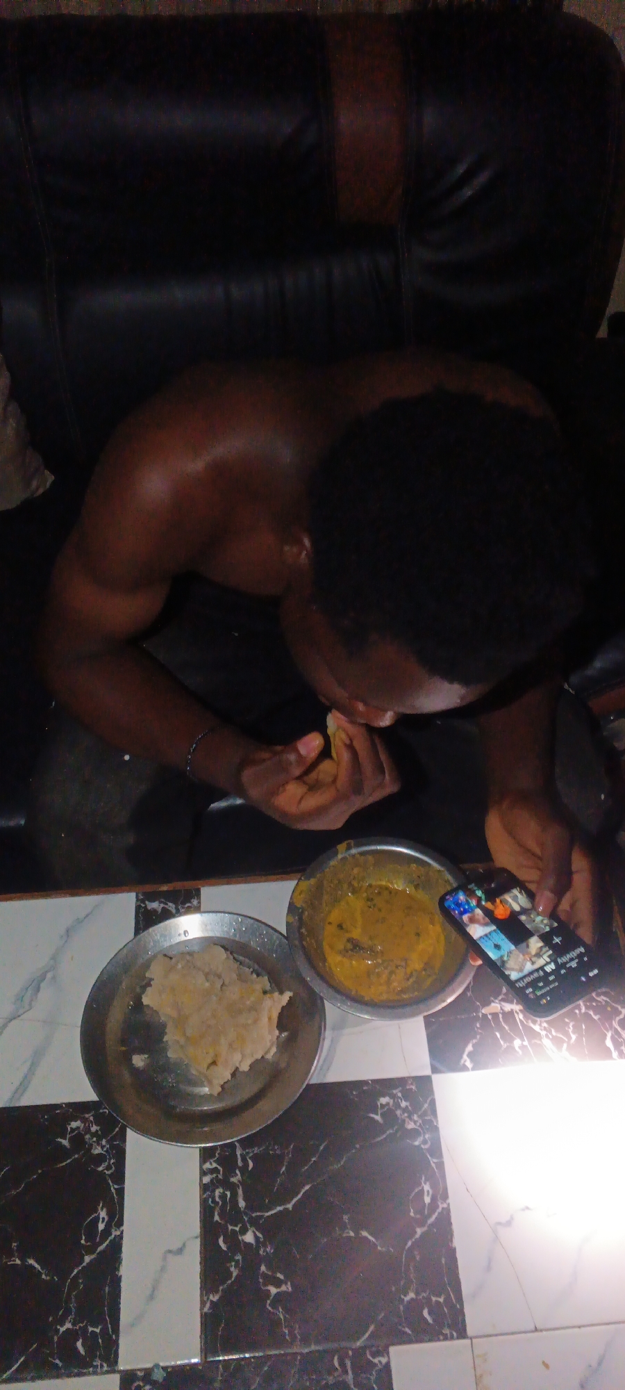 A dark-skinned man with short black hair sits shirtless on a black leather couch. He is eating from a metal bowl containing yellow soup with visible pieces of something, possibly meat or vegetables. Next to it is a plate of what appears to be "swallow" or a similar starchy food. He is looking at his phone with his left hand as he eats. The phone screen displays various images in a grid, possibly on a social media app. The scene is indoors. The table has a black-and-white tiled pattern. The exact location in Onitsha, Nigeria is not discernible.