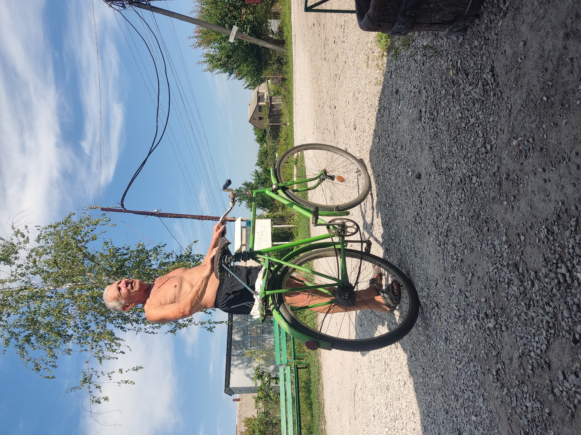 An elderly man with a bare torso and dark shorts stands next to a green bicycle on a gravel road in Radisne, Ukraine. He is facing to the right, looking upwards. The bicycle is positioned at an angle, with its front wheel slightly ahead of the man's feet. Behind the man, a large tree with sparse leaves is visible, along with utility poles and power lines against a blue sky with scattered clouds. To the left, a green park bench sits in front of a building. The ground is covered in light-colored gravel. The overall scene suggests a rural or suburban setting on a sunny day.