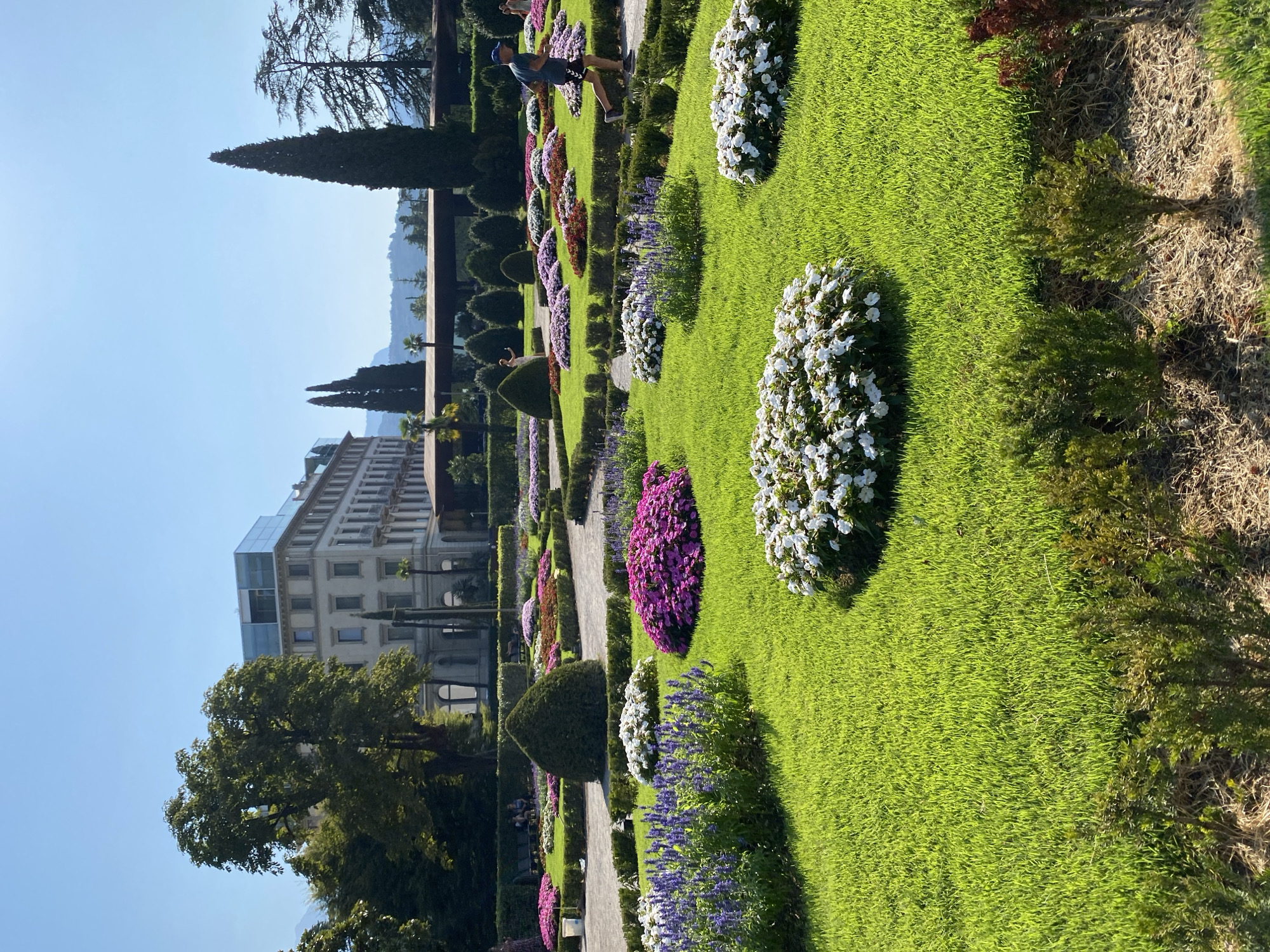 The image shows a meticulously landscaped garden in front of a grand building, with a person jogging in the foreground. The garden features a vibrant lawn, manicured hedges, and various flowerbeds displaying an array of colorful blooms, including purple, pink, and white flowers. In the background, a large, multi-story building with a modern glass-encased top floor stands prominently. Tall, slender cypress trees and other greenery dot the landscape, with hills and mountains visible in the distance under a clear blue sky. This scene is located at the Grand Hotel Riva, situated in Riva del Garda, Italy. The person jogging is a male wearing a blue cap, dark t-shirt, and black shorts, running on a paved path within the garden. Other individuals can be seen seated on benches in the background, enjoying the garden. The environment is sunny and appears to be a pleasant day.
