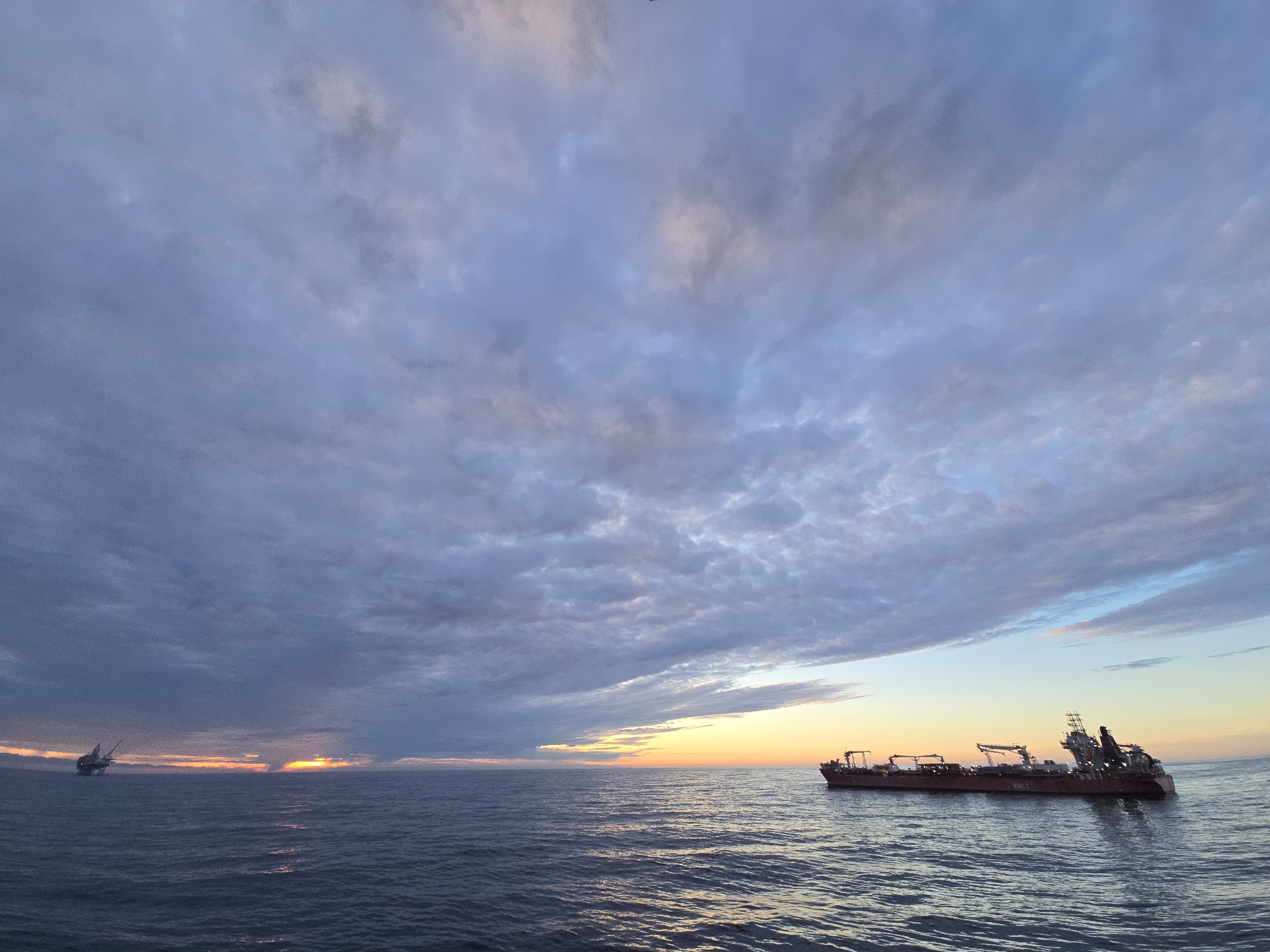 The image captures a seascape at sunset under a sky filled with textured clouds. To the left, a partially visible offshore oil platform stands against the horizon where the sun is setting. To the right, a red barge-like vessel is seen on the water. The location is Unknown, Unknown.