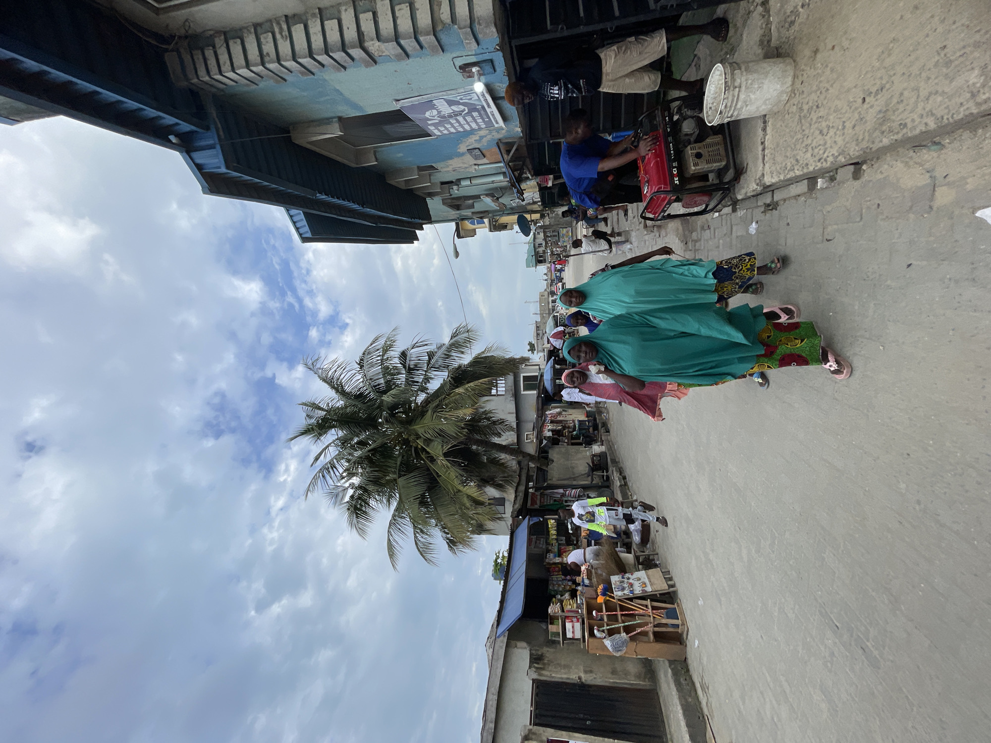 The image shows a street scene in Lekki, Nigeria. Several people walk along the street, including a group of women wearing green hijabs. A coconut tree stands prominently. A man is operating a small gasoline-powered generator set on the sidewalk. There are shops and vendors with displayed goods along the street. The sky is cloudy.