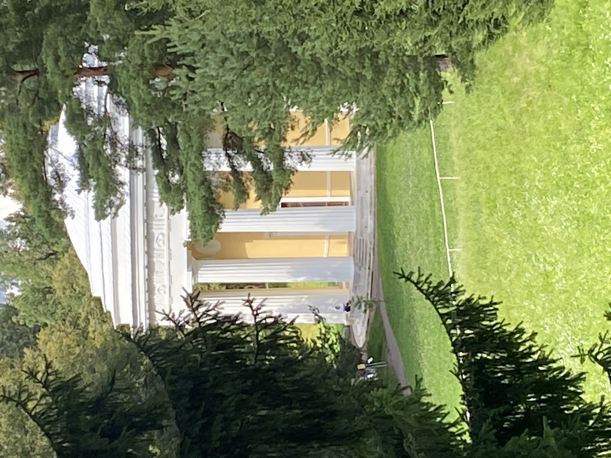 The image depicts a neoclassical pavilion with a white roof and yellow walls, framed by dark green coniferous trees. The pavilion is supported by a row of white columns, and stairs lead up to its entrance. A person in a white top and dark pants is visible sitting on the stairs. The pavilion is situated in a park with a well-maintained green lawn and a pathway winding through it. In the background, a line of trees forms a dense backdrop. This scene is characteristic of the Pavlovsk Park, a sprawling landscape park located in the town of Pavlovsk, Pushkinsky District, Saint Petersburg, Russia.