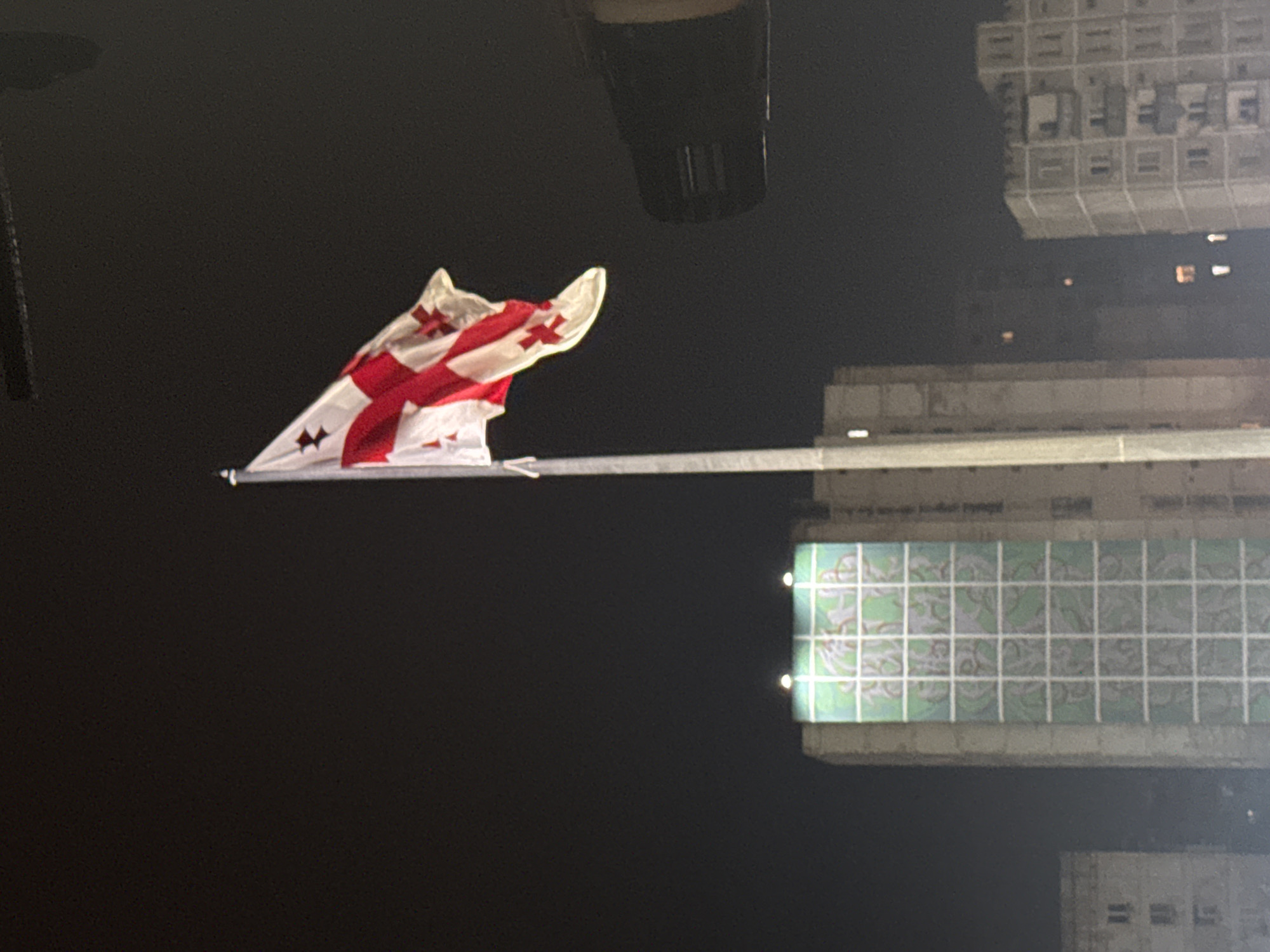 The image depicts a Georgian flag on a flagpole at night, with buildings in the background. The flag, predominantly white with a red cross and four smaller red crosses, is illuminated and appears to be waving. To the left of the flagpole, a tall, rectangular building features a large, green mural with white grid lines. To the right of the flag, a portion of a dark, cylindrical object, possibly a light fixture or a camera, is visible. Further in the background, several other buildings are visible, some with illuminated windows. The scene is set in an urban environment at night, with the dark sky providing a backdrop. Based on the distinctive architecture of the buildings and the prominent display of the Georgian flag, this image is likely taken in Tbilisi, Georgia.