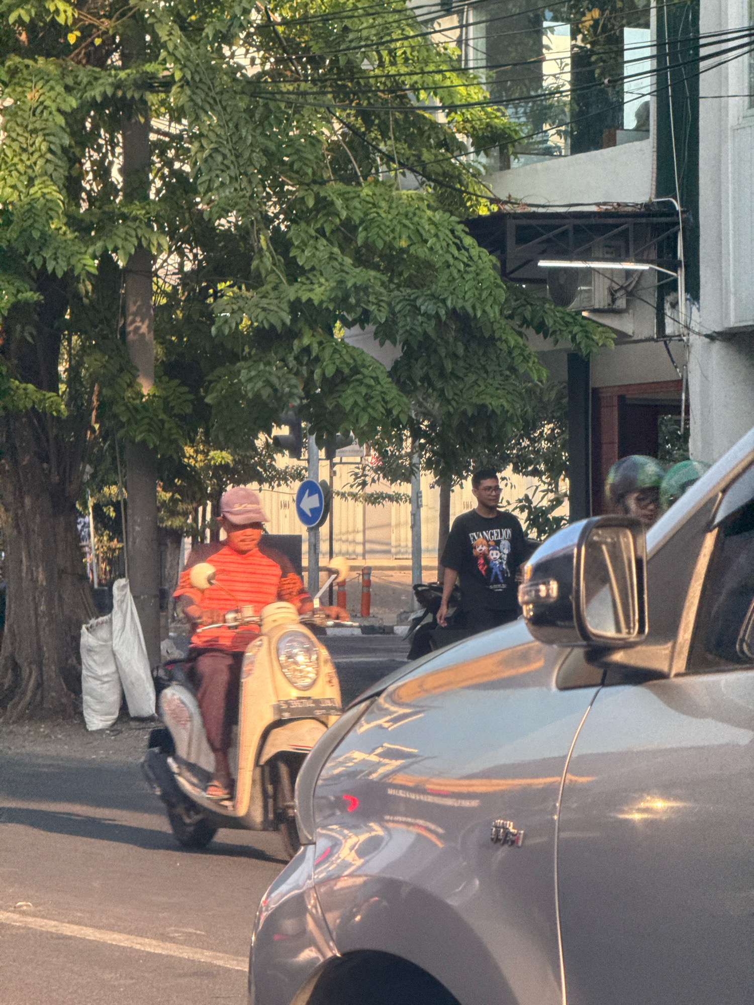 The image depicts a street scene in Surabaya, Indonesia, during daylight. In the foreground, the right side of a silver car is visible, with its side mirror and part of its fender. To the left of the car, a person is riding a light-colored scooter. This individual is wearing a reddish-orange striped long-sleeved shirt and dark pants, with a light-colored cap. Behind the scooter rider, a young man in a black t-shirt with graphic print is standing on the sidewalk. Further behind him, two individuals wearing green helmets are visible, one of whom appears to be riding a motorcycle. The background features lush green trees, a building with reflective windows, and utility wires. A traffic sign with a blue circular background and a white arrow pointing left is also present. The overall lighting suggests it is either early morning or late afternoon, with the sun casting long shadows.