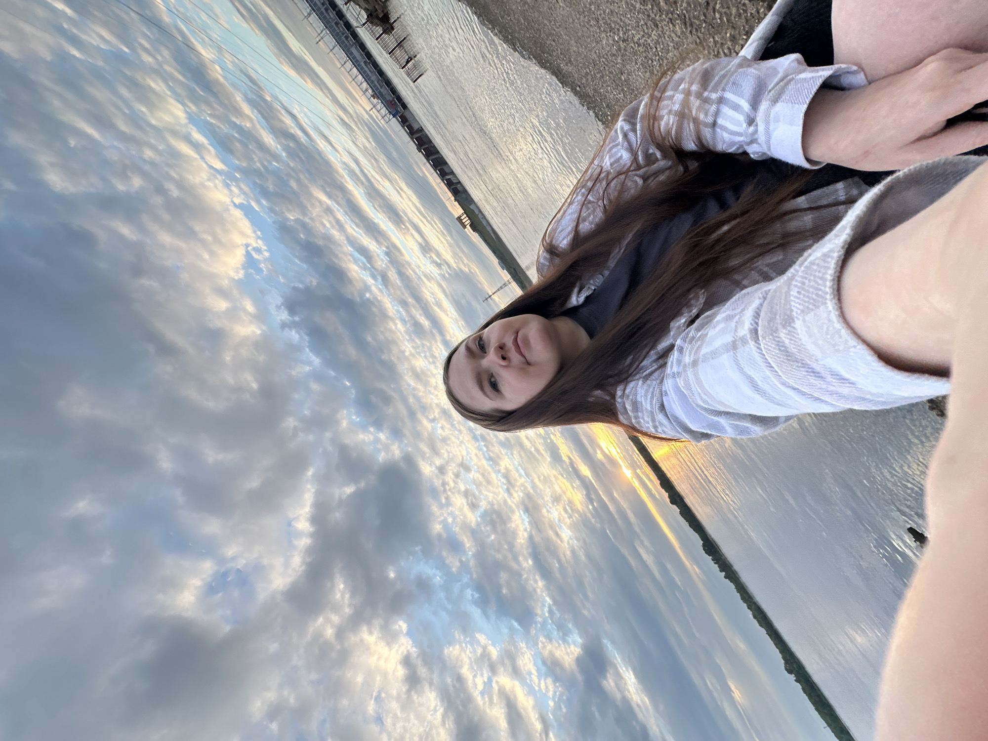 A young woman with long, brown hair is seated outdoors on a gravelly shoreline, looking directly at the camera. She is wearing a light-colored, plaid button-up shirt over a dark t-shirt. The background features a wide body of water reflecting the sky. The sky is filled with dramatic, scattered clouds tinged with the colors of sunset, with a hint of sunlight peeking through on the horizon. In the distance, across the water, a large dam structure and industrial buildings are visible.