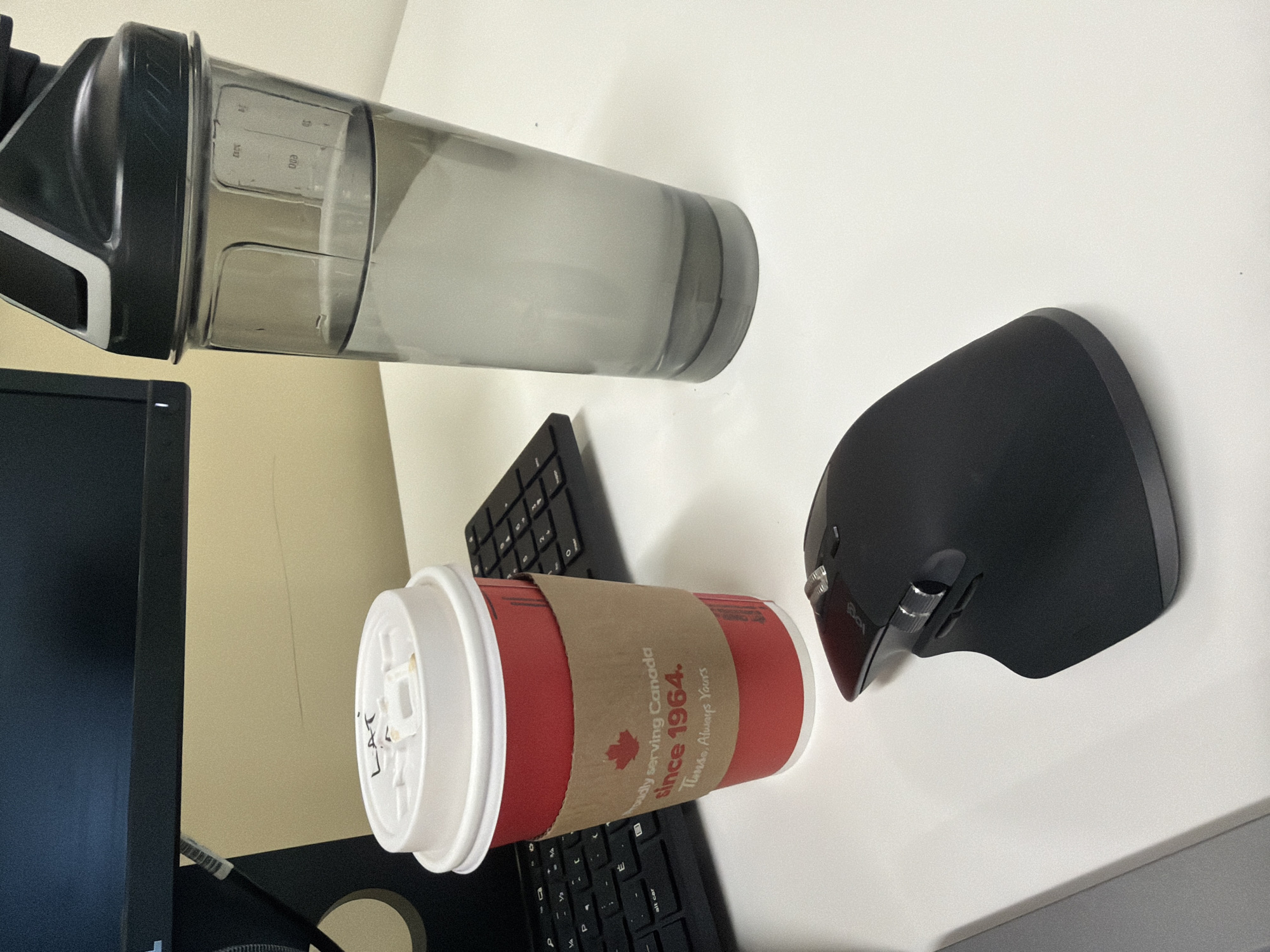 This image depicts a static indoor scene on a white desk, likely within an office setting in Mirabel, Canada. Positioned centrally in the foreground is a red Tim Hortons disposable coffee cup with a white lid. The lid has "LAT" handwritten in black marker. A brown cardboard sleeve around the cup displays a red Canadian maple leaf logo and the text "Proudly serving Canada since 1964. Always Yours." To the right of the cup stands a grey, translucent shaker bottle, approximately half-filled with clear liquid, presumably water, exhibiting visible measurement markings on its side. A black ergonomic computer mouse with multiple buttons and scroll wheels, bearing a "logi" brand inscription, is situated in front of the coffee cup. Partially visible to the left and behind the coffee cup is a black computer keyboard, showing a QWERTY layout with French Canadian keycaps. A dark-colored computer monitor screen and its lower bezel are visible in the background, above the desk, against a plain light beige wall. No individuals are present in the frame, and no actions or direct interactions are occurring.
