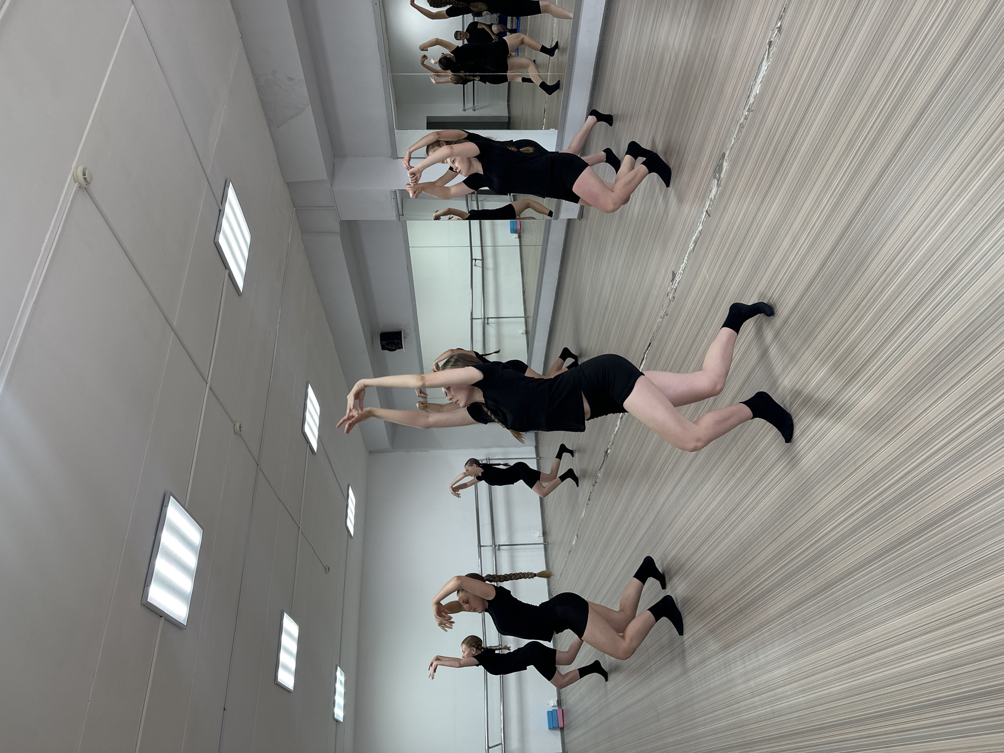 The image shows a dance studio with several young women in black athletic wear performing a choreographed dance. They are barefoot, wearing black socks. The studio has a wooden floor, a mirrored wall along one side, and a ballet barre. Fluorescent lighting fixtures are visible on the ceiling. The dance studio is located in Votkinsk, Russia.