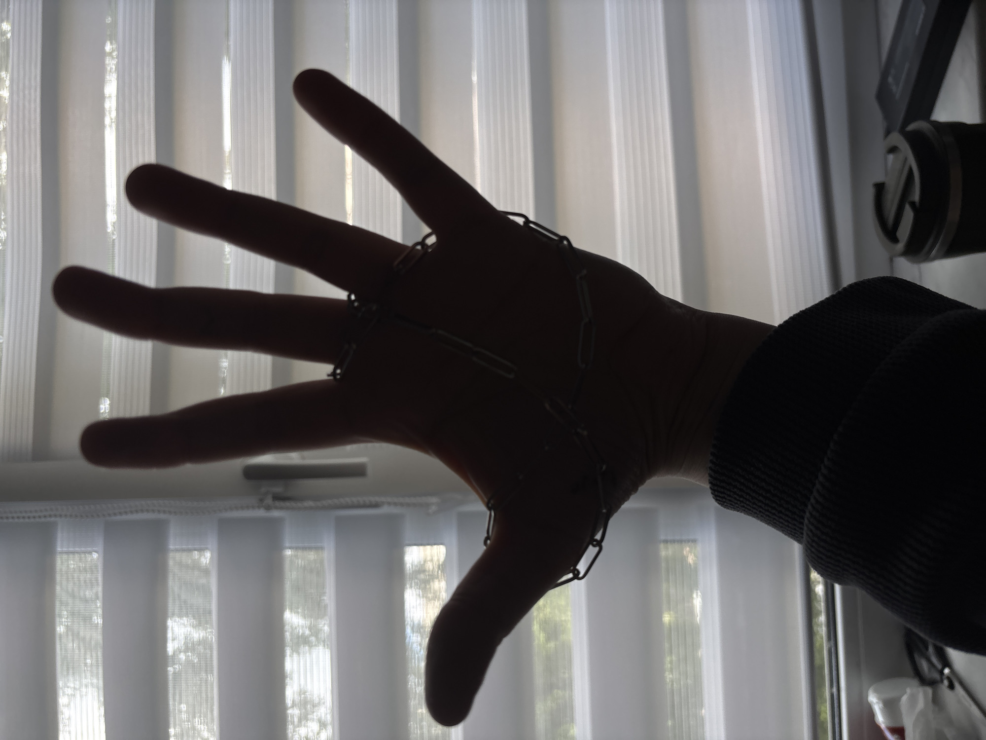A silhouette of a hand is the main subject of this image. The hand, with fingers spread, is adorned with a chain that wraps around multiple fingers. The hand is positioned in front of vertical blinds that allow diffuse light to pass through, suggesting it might be daytime. The light filtering through the blinds creates a soft, textured background. In the upper right corner, a portion of a camera or microphone is visible, also in silhouette, adding to the enigmatic feel of the image. The overall mood is somewhat mysterious due to the strong contrast and the focus on the isolated hand and chain. The setting appears to be indoors, near a window with blinds. There is no visible text in the image. The location context is Saint Petersburg, Russia.