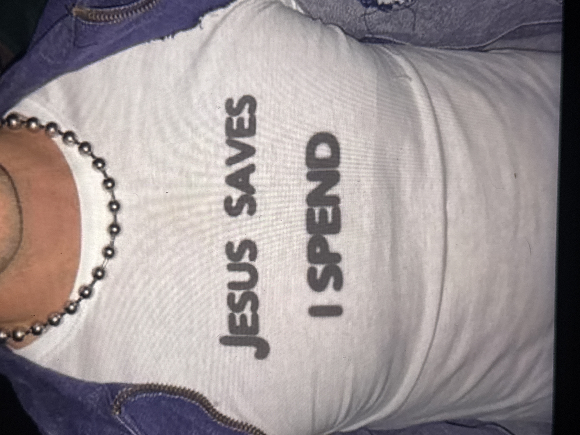 The image is a close-up, vertically oriented shot of a person wearing a white t-shirt with dark text that reads "JESUS SAVES I SPEND". The person is also wearing a silver beaded necklace and a blue denim jacket that is partially visible at the top and bottom of the frame. The lighting is dim, and the focus is on the t-shirt. The setting appears to be indoors, possibly a casual environment. There are no clear indicators of time of day, weather, or specific location cues, other than the provided location context of Kuta Selatan, Indonesia. The overall emotion conveyed is neutral, as the focus is on the text and clothing.