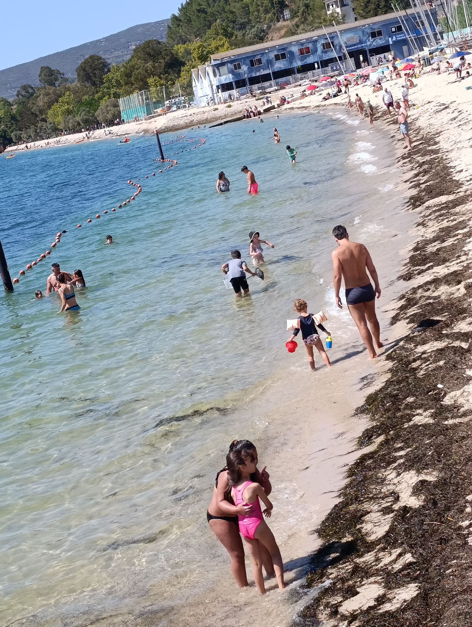 The image depicts a sunny day at a beach in Setúbal, Portugal, likely the Praia da Saúde due to the presence of the nautical club building in the background. The water is clear and shallow, with visible seaweed on the sandy bottom. Numerous people are enjoying the beach and the water. In the foreground, two women, one adult and one child, are partially submerged in the water, with the child wearing a pink swimsuit. To their right, a man in dark swim trunks is standing on the wet sand, facing away from the camera. Further into the water, a family group consisting of a man, two women, and two children are interacting. Other individuals are scattered across the beach and in the water, engaged in various activities such as swimming and playing. The background features a building with blue accents, identified as a nautical club, with sails visible on the property. A wooded hillside rises behind the beach.