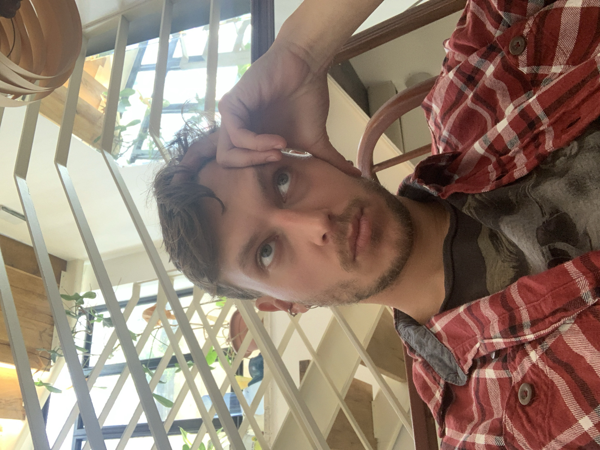 A medium close-up, rotated portrait shows a man with light brown hair and stubble, wearing a red plaid shirt over a darker t-shirt. His right hand is raised to his forehead, with a round object visible on his fingers. The man has a serious expression, looking directly at the camera. Behind him is a latticework structure with plants visible through the gaps, suggesting an indoor garden or greenhouse setting. A wooden chair is partially visible on the right. The scene has a bright, natural lighting. There is no concrete evidence that the location is Tel-Aviv, Israel.