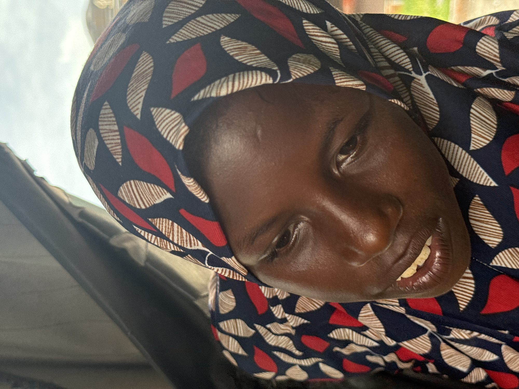 A close-up, slightly tilted portrait captures an individual with dark skin, likely a woman, wearing a patterned head covering. The subject's face is prominent, with their gaze directed towards the viewer, and a gentle smile or smirk playing on their lips, revealing a glimpse of teeth.
The head covering is made from fabric featuring a distinct pattern of dark blue (or black) as the base, adorned with repeating leaf-like or abstract shapes in vibrant red, white, and light brown. The white and light brown shapes are further detailed with fine horizontal stripes.
The lighting is bright and natural, indicating it is daytime. The background suggests an outdoor or open-air setting, with a bright, overcast or cloudy sky visible in the upper left portion of the image, and darker, ambiguous shapes (possibly a structure or vehicle interior) in the lower left.
Notable details include a natural sheen on the person's skin, a subtle mark or scar on the forehead, and the intricate design of the head covering. The expression conveys a sense of quiet warmth. There is no visible text in the image. The setting is consistent with a casual moment captured in a location like Ado, Nigeria.