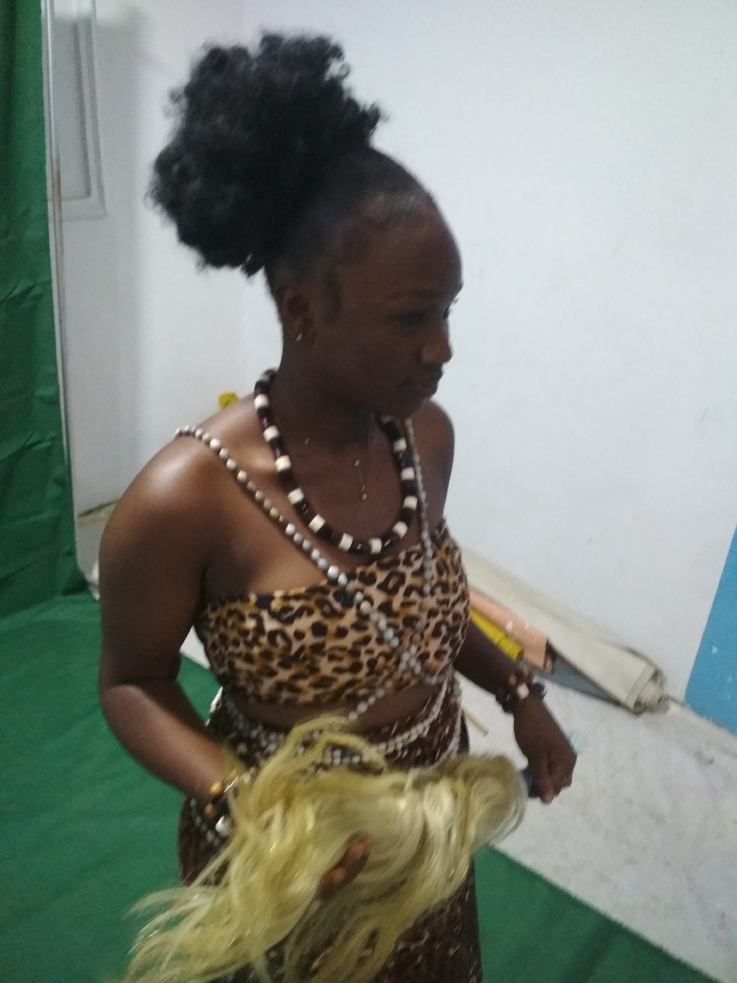 A woman is depicted indoors, possibly preparing for a performance or event. She has dark skin, wears her hair in a high bun, and dons jewelry, including a two-strand beaded necklace and a bracelet. Her attire consists of a leopard-print top and a matching skirt. She is holding what appears to be light-colored hair extensions or a wig in her hands. The background includes a green cloth or screen to the left, white walls, and various objects suggesting a dressing room or preparation area. The specific location within the city of Unknown, Unknown, is not discernible.
