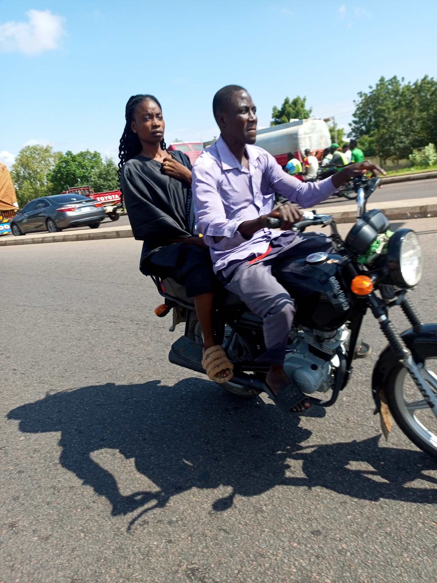 In Tempure, Nigeria, a man and a woman ride a black motorcycle on a paved road under a clear blue sky. The man, wearing a light purple collared shirt and dark pants, drives the motorcycle. The woman, with braided hair, is seated behind him wearing a black top and dark pants. Both are casting long shadows on the asphalt. In the background, other vehicles, including a gray sedan with the "TOYOTA" logo visible on a red truck, and trees are present, indicating a roadside environment. The motorcycle appears to be in motion.