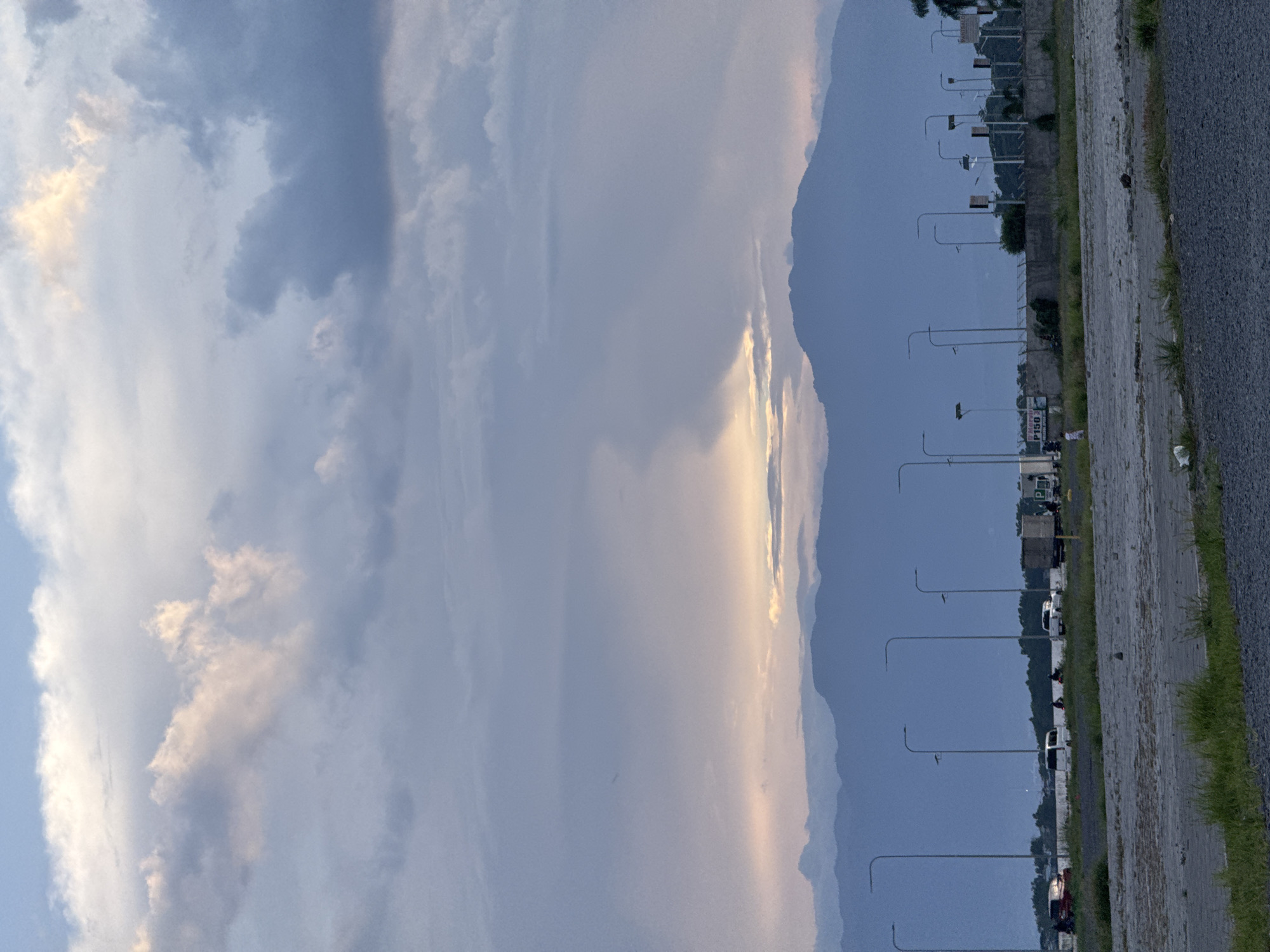 The image shows a sideways view of a road near the mountains in Davao City, Philippines. The road runs along the right side of the image with street lights evenly spaced, and a number of parked vehicles are visible. Mountains are in the background next to the road. The sky is cloudy, with some patches of light suggesting the sunset.