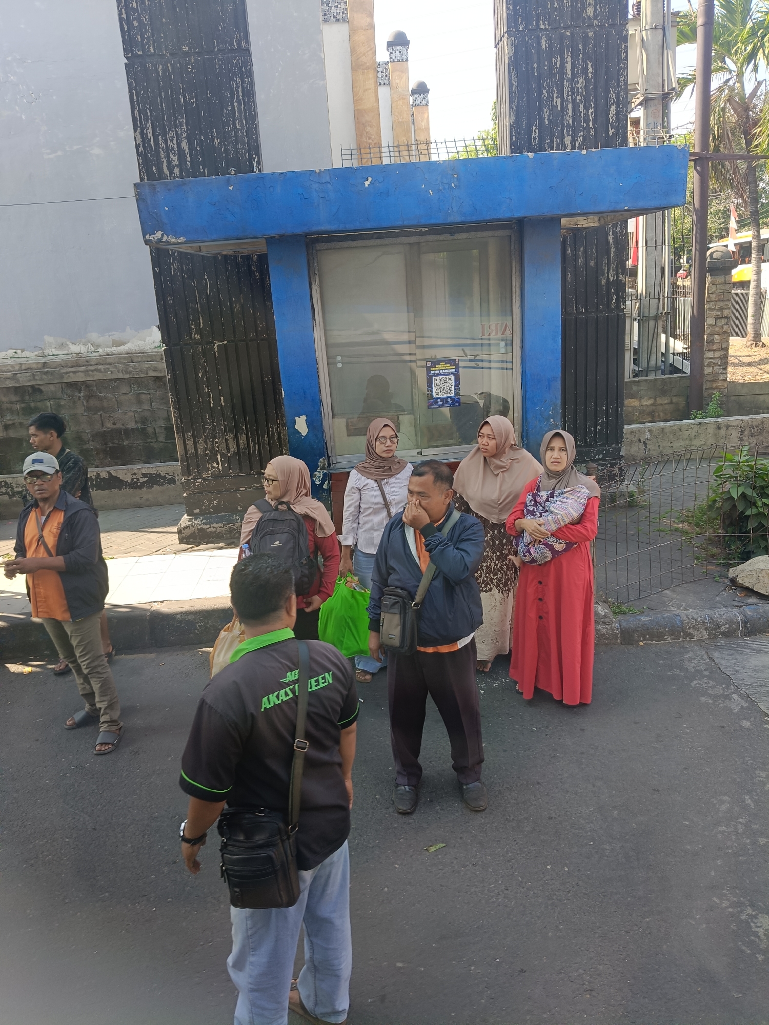 A group of seven people, five women and two men, are standing on a paved street in front of a building with a blue facade and large black pillars. The building appears to be a storefront or public facility. The people are dressed casually, with several of the women wearing headscarves. One woman in the foreground is holding a baby wrapped in a colorful cloth. Another man is wearing a black t-shirt with green lettering that reads "AKAST IJEEN." The environment suggests an urban setting in Kedungrejo, Indonesia, with a sidewalk, a metal fence, and some greenery visible in the background. The lighting indicates it is daytime, likely morning or afternoon, given the shadows.