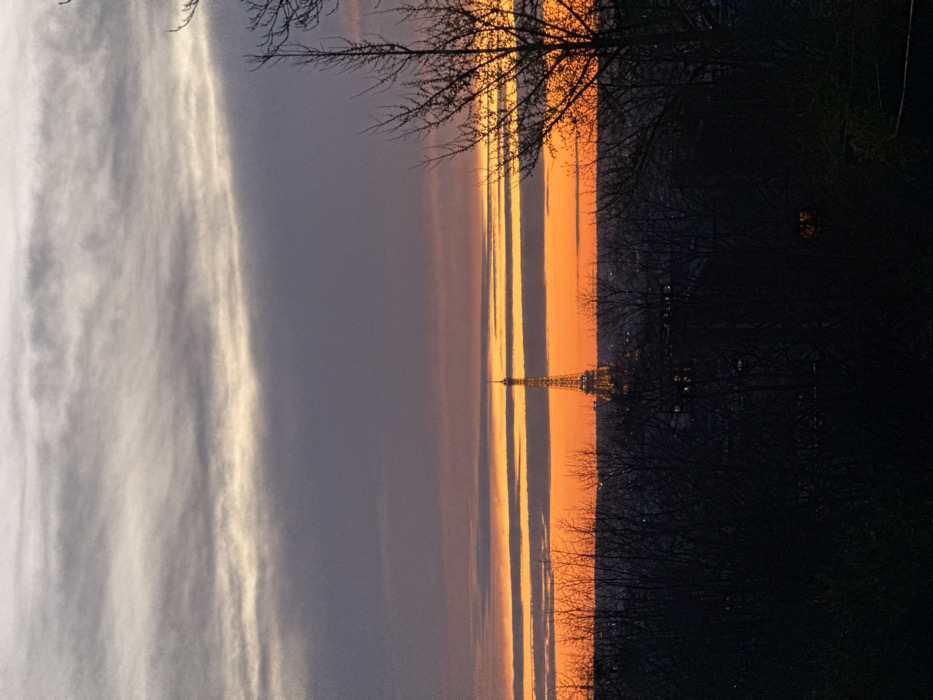 The image captures a panoramic view of Paris, France, during twilight, likely sunset, from an elevated vantage point looking west. The sky dominates the upper two-thirds of the frame, featuring layered clouds transitioning from light grey and white at the top, through a wide band of muted purplish-grey, to a prominent horizon displaying vibrant orange and red hues with distinct horizontal cloud formations.
The Eiffel Tower, illuminated with a warm yellowish light, stands centrally in the mid-ground, silhouetted against the bright orange portion of the sky. Its characteristic lattice structure is visible. Below the horizon line, the general silhouette of the Parisian cityscape is faintly discernible between foreground elements, with a few small lights visible within the urban sprawl.
The foreground and lower mid-ground are occupied by dense, dark silhouettes of numerous deciduous trees, appearing bare of leaves, indicating either a winter or early spring season. These trees partially obscure the lower sections of the city. No individuals or overt actions are depicted. The scene suggests an observation point from an eastern district of Paris, such as Montmartre or Parc de Belleville.