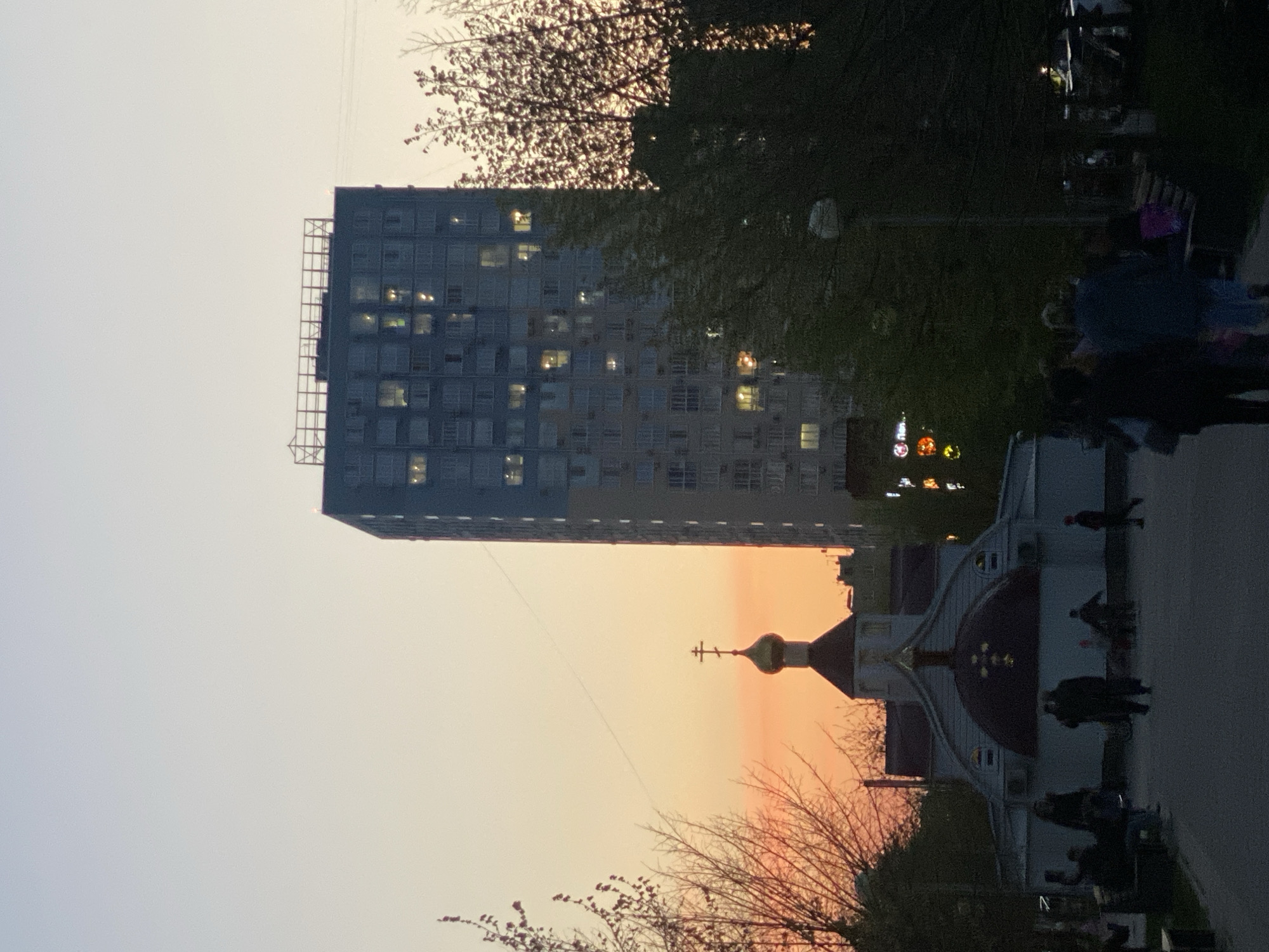 The image captures a street scene at dusk in an urban location. Dominating the background is a high-rise building with multiple windows illuminated, suggesting evening. In the foreground, there is an orthodox church structure with a distinctive dome and a cross on top. People are present on the pathway near the church, appearing as silhouettes. Trees, some bare, frame the scene, adding to the urban-nature mix. The sky exhibits a gradient of colors from lighter tones to orange hues, indicative of sunset or twilight.