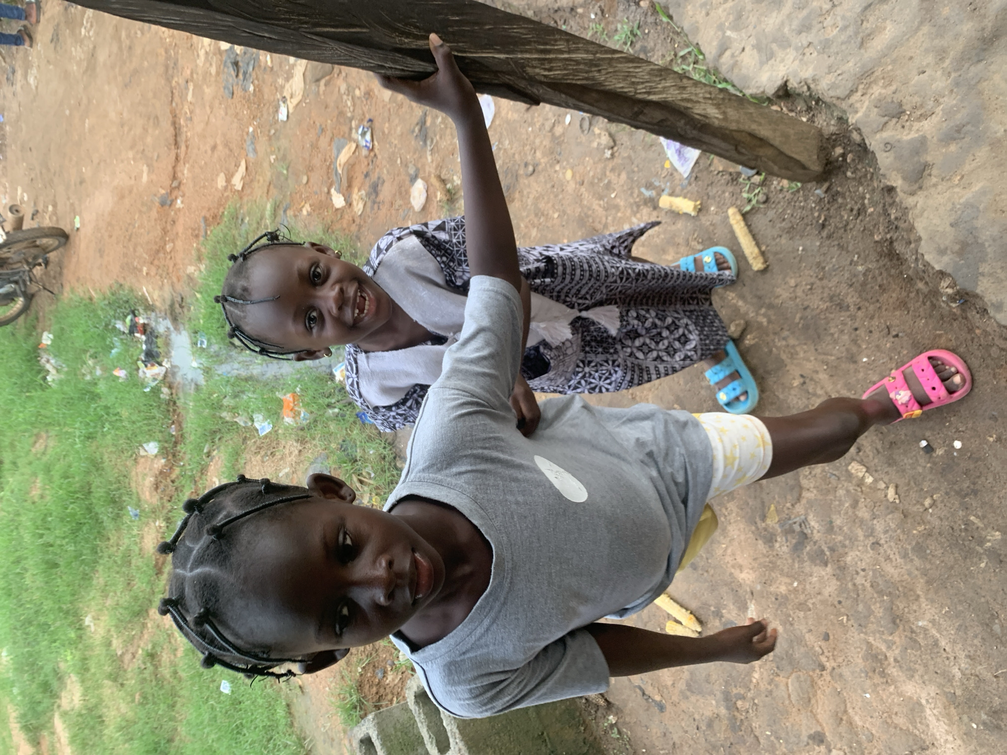 The image shows two young Black children standing outdoors on a dirt path in Shagamu, Nigeria. The child on the left, appearing to be older, is wearing a grey t-shirt and yellow shorts with a star pattern. The child on the right, who is smiling broadly, is wearing a black and white patterned dress and blue sandals. The ground is rough and uneven, with scattered debris including plastic bags and what appears to be corn cobs. In the background, there is a grassy area with more scattered trash and a glimpse of a motorcycle. The overall scene suggests a humble, possibly impoverished, urban or semi-urban environment.