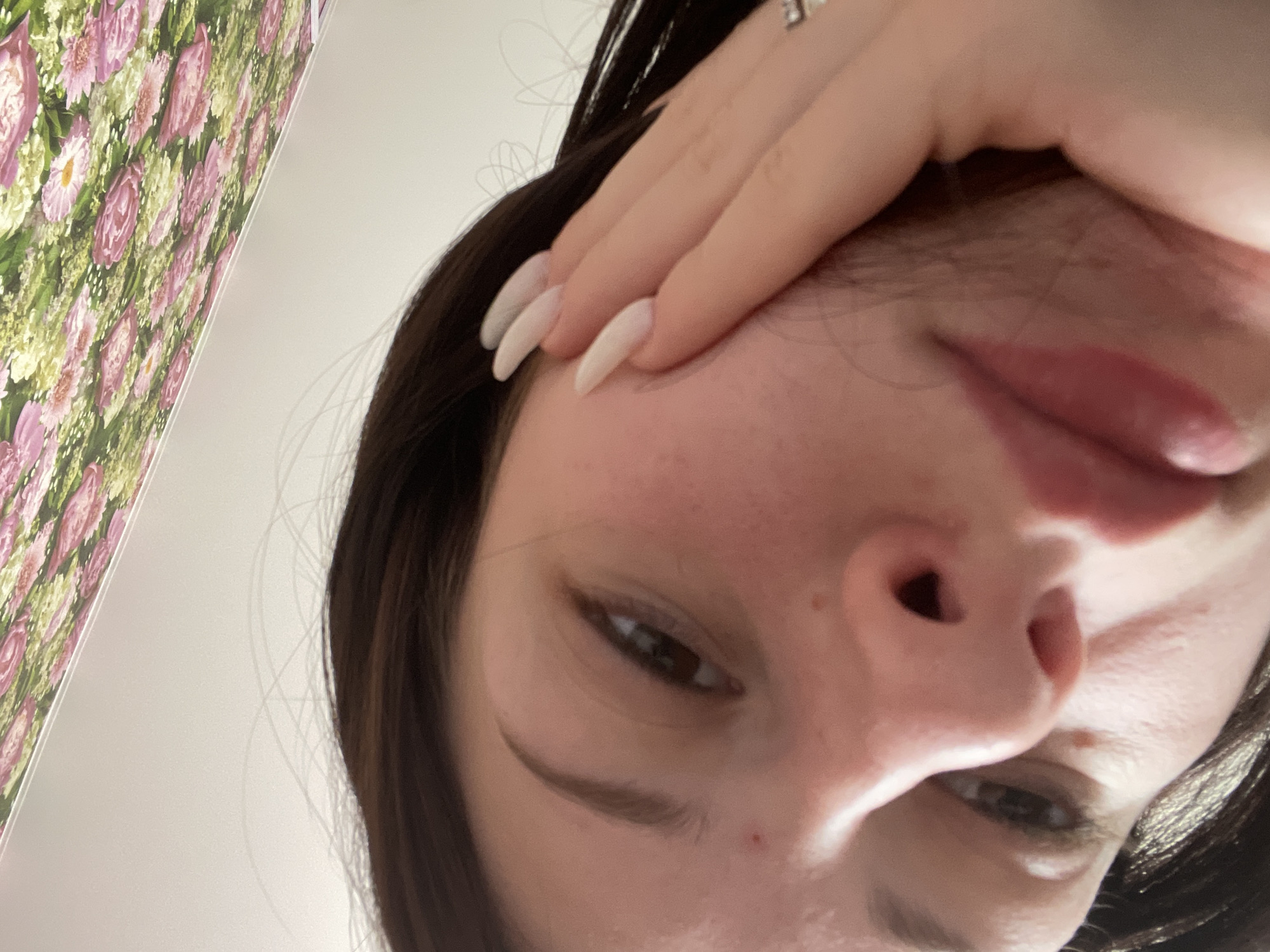 A close-up, low-angle portrait features a young individual, situated in Медведево, Russia. The subject's face is central, with their dark hair falling around it. One hand, adorned with long, pale, coffin-shaped nails and a silver-toned ring on a finger, rests against their forehead, partially obscuring the upper part of their face. The individual looks towards the camera with dark eyes, and their mouth is slightly ajar, revealing the tip of their tongue.
To the upper left, a vibrant floral pattern, possibly wallpaper or fabric, with distinct pink and purple flowers amidst green and light-colored foliage, frames a portion of the scene. The rest of the background consists of a plain, light-colored wall or ceiling. The lighting is even and soft, suggesting an indoor setting. The image captures a casual, intimate moment, with no visible text.