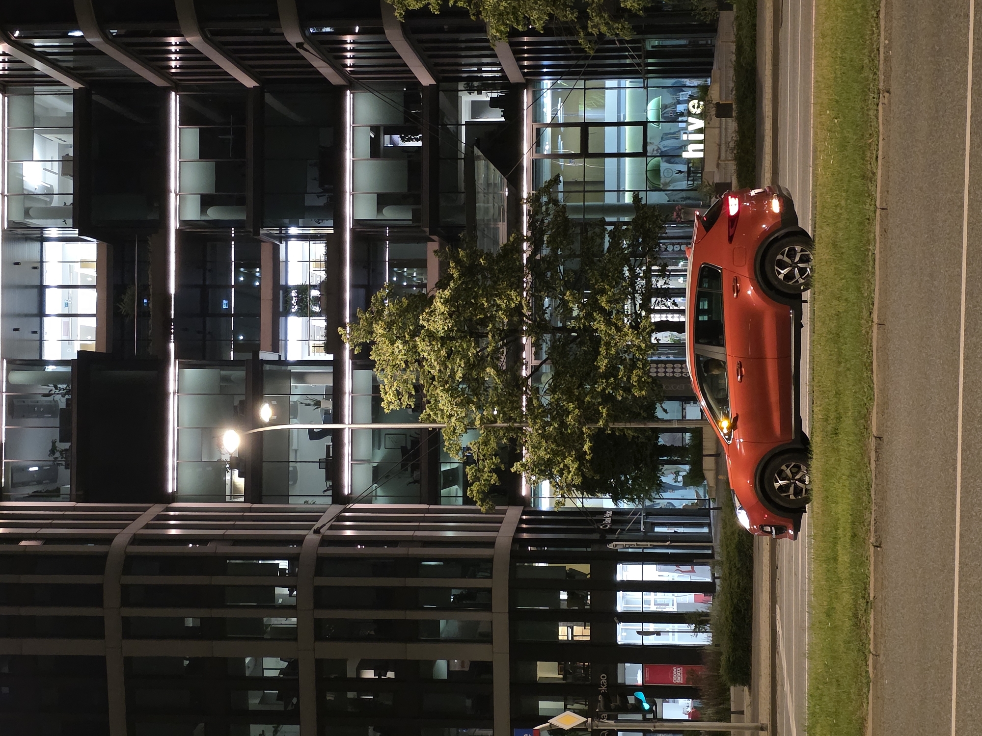 An orange Kia Sportage is parked on the side of a road at night, with a modern office building in the background. The office building has large glass windows that are lit up from within, revealing the interiors of several floors. A large, leafy tree is positioned in front of the building, partially obscuring some of the windows. The building has a contemporary architectural style with multiple levels and protruding sections. The street in front of the building appears to be a multi-lane road with a grassy median separating the lanes. There are streetlights illuminating the scene. The building has a prominent sign that reads "hive". The location is identified as Warsaw, Poland.