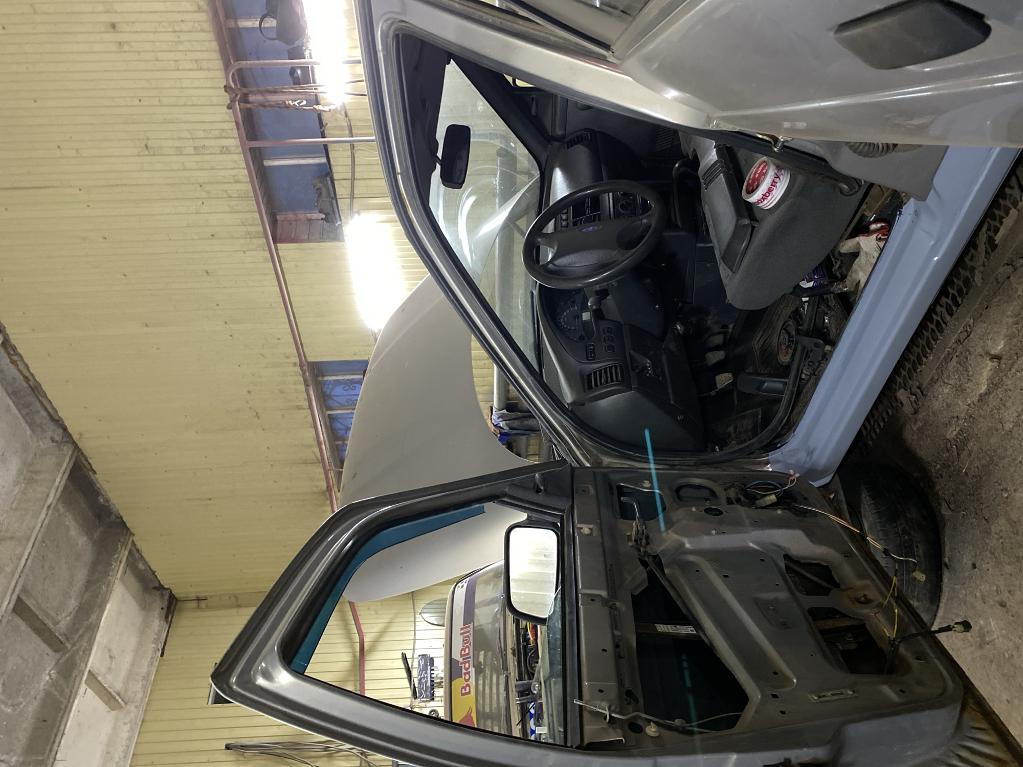 The image shows the interior of a vehicle undergoing repair or restoration, likely in a garage or workshop in Chaikovsky, Russia. The vehicle, possibly a van or truck, has both its hood and driver's side doors open. The driver's side door is stripped down to its metal frame with visible wiring. The interior is partially disassembled, exposing parts of the dashboard, steering wheel, and seats. There is a can with "oxberry" on the lid, a brand that is made in Russia, situated near the seat. A "RedBull" sign is visible through the window on the opened door. The garage or workshop is characterized by its exposed walls and ceiling with overhead lighting. No people are visible in the image.
