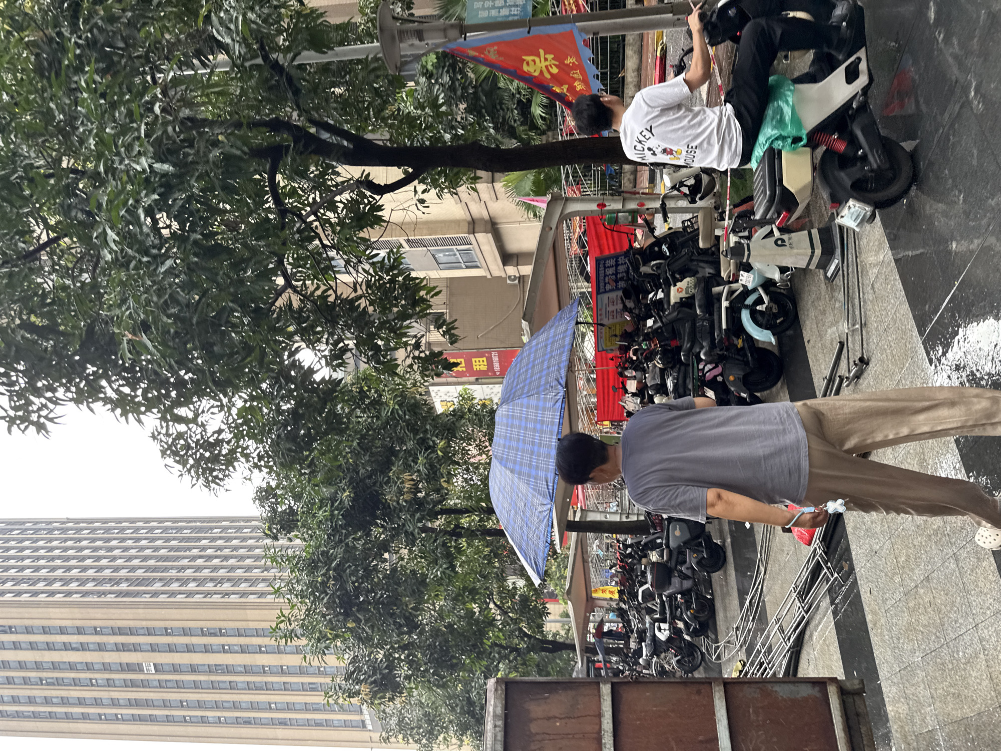 In the Haizhu District of China, a city scene unfolds under an overcast sky, possibly after rain, as evidenced by the wet pavement. In the foreground, a person wearing a grey t-shirt and beige pants walks away from the camera, holding a small blue and white charm. Behind them, a large blue and white checkered umbrella is propped up, providing shelter. A crowd of electric scooters and motorcycles are parked in rows under awnings. To the right, a person in a white t-shirt with a Mickey Mouse graphic is seated on a scooter, facing to the right and out of frame. A red and yellow triangular flag hangs from a pole nearby. In the background, a tall, modern building with many windows stands prominently, surrounded by trees. The overall atmosphere suggests an urban environment, likely a public space where transportation is readily available.