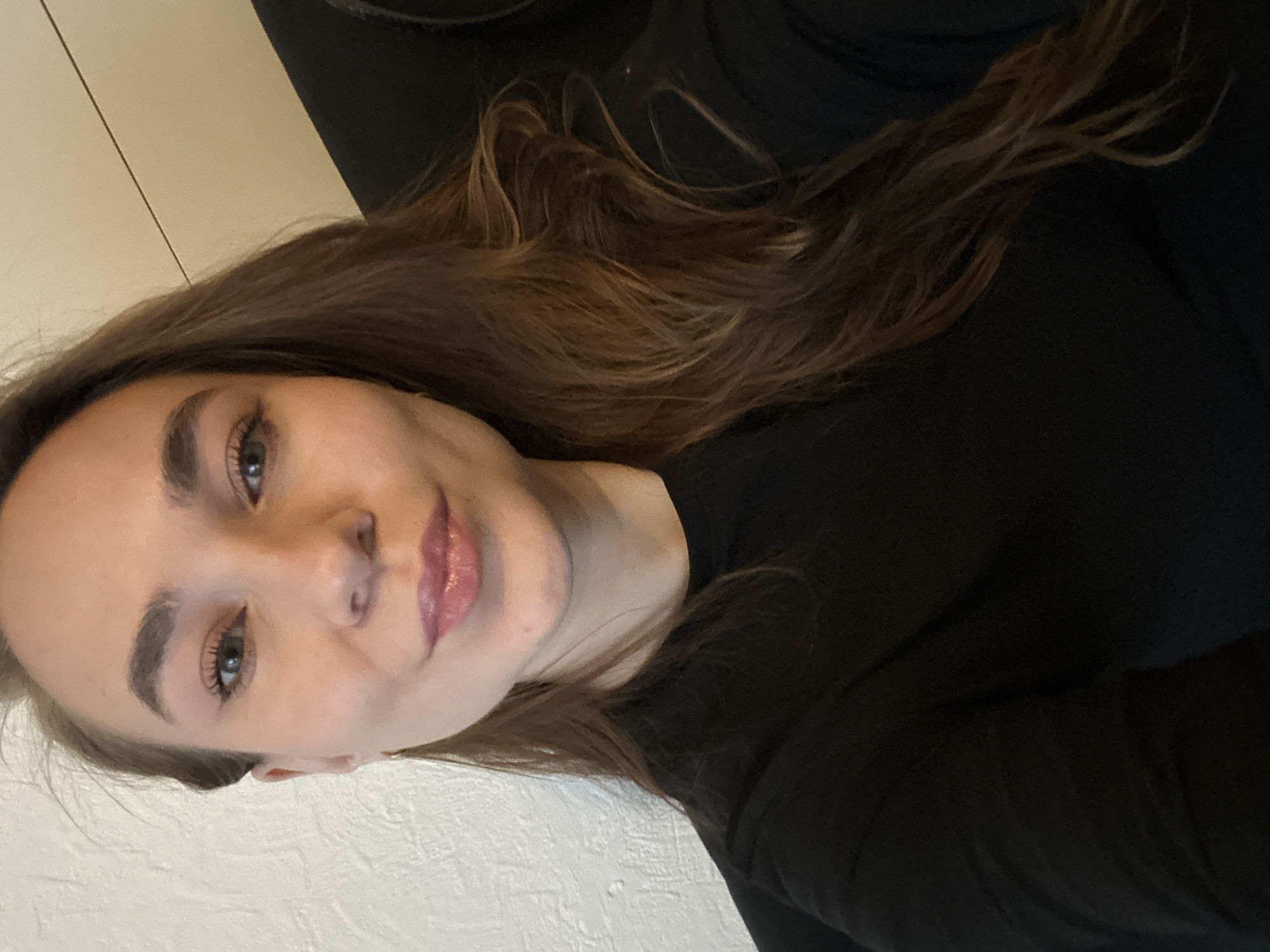 The image shows a selfie of a young woman with light skin, brown hair, and hazel eyes. She is wearing a black long-sleeved shirt. The background appears to be a textured white wall on the left. The location in the city of Chaikovsky, Russia is not discernible from the image.